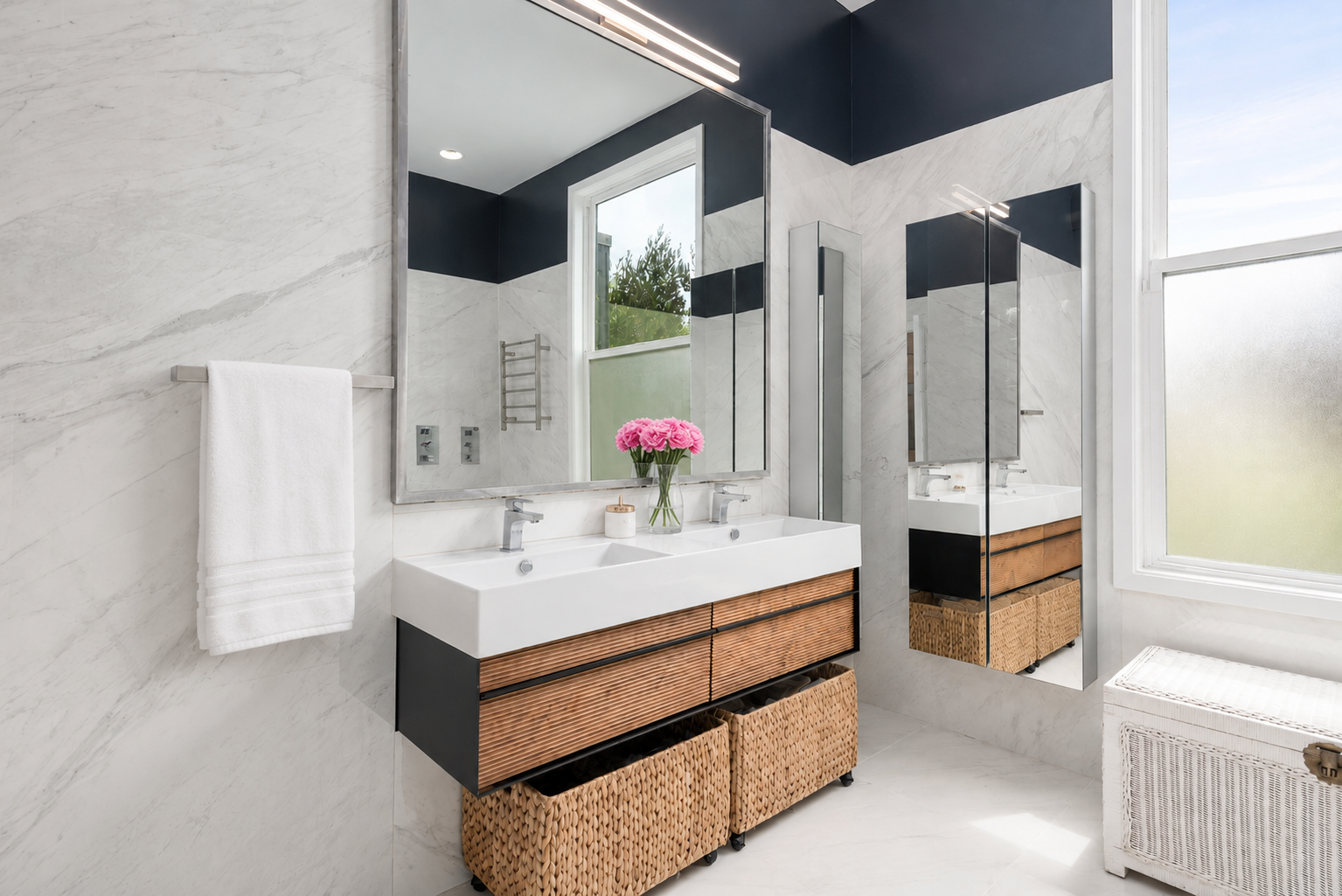Modern bathroom with double vanity, large mirror, two sinks, a pink flower bouquet, wicker storage baskets, and a window with frosted glass.