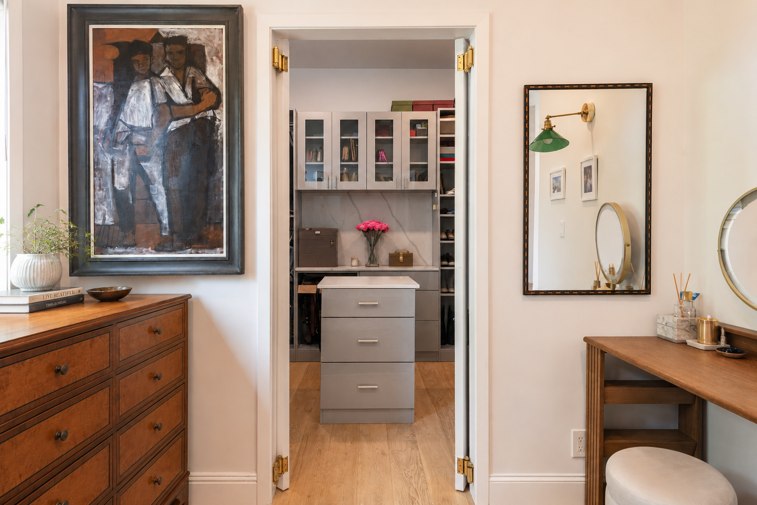 A walk-in closet with a gray dresser and glass-front cabinets, pink flowers on top, and shoes on shelves. The closet leads to a room with wooden furniture and decorated walls.