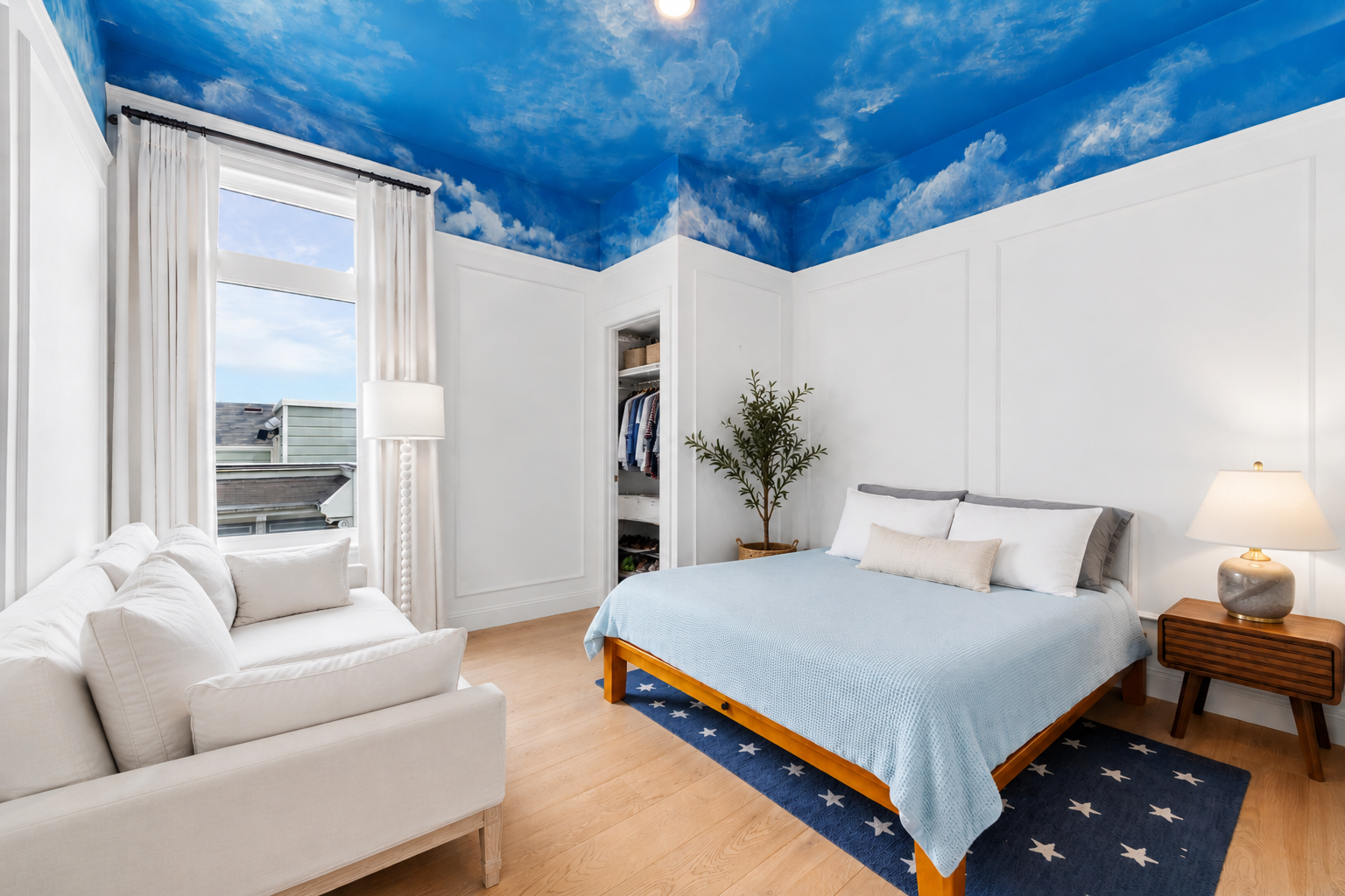 A bedroom with a blue sky cloud-themed ceiling, white walls with wall paneling, a window with white curtains, a white couch, a bed with a wooden frame, gray and white pillows, a wooden nightstand with a lamp, a potted plant, and an open closet with c