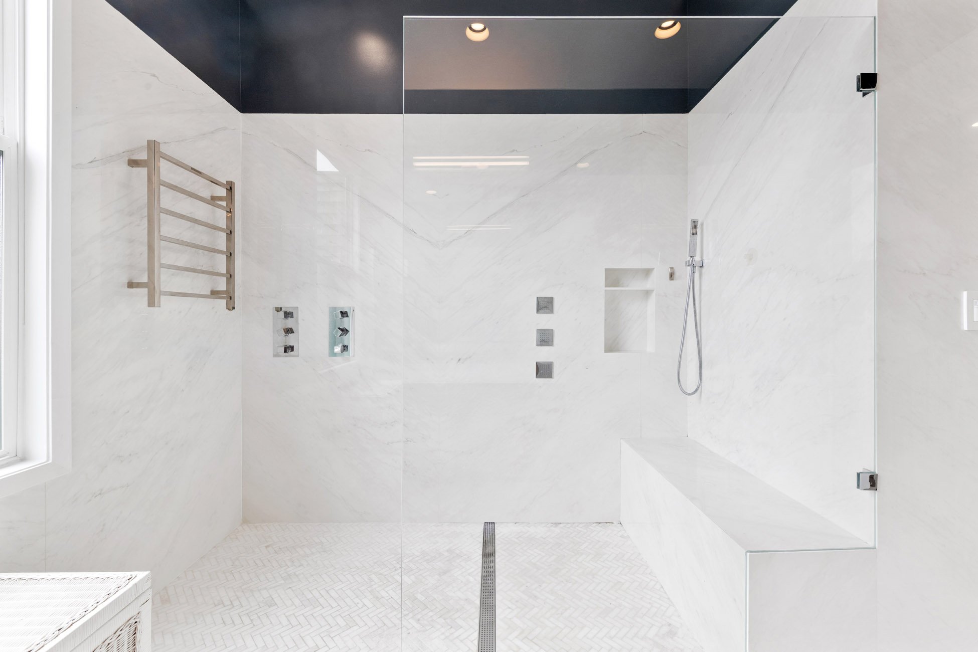 Modern walk-in shower with white marble walls and a black ceiling, featuring multiple showerheads and a handheld shower. There is a wooden towel rack on the left wall and built-in shelves in the shower area. The shower has a glass enclosure and a lin