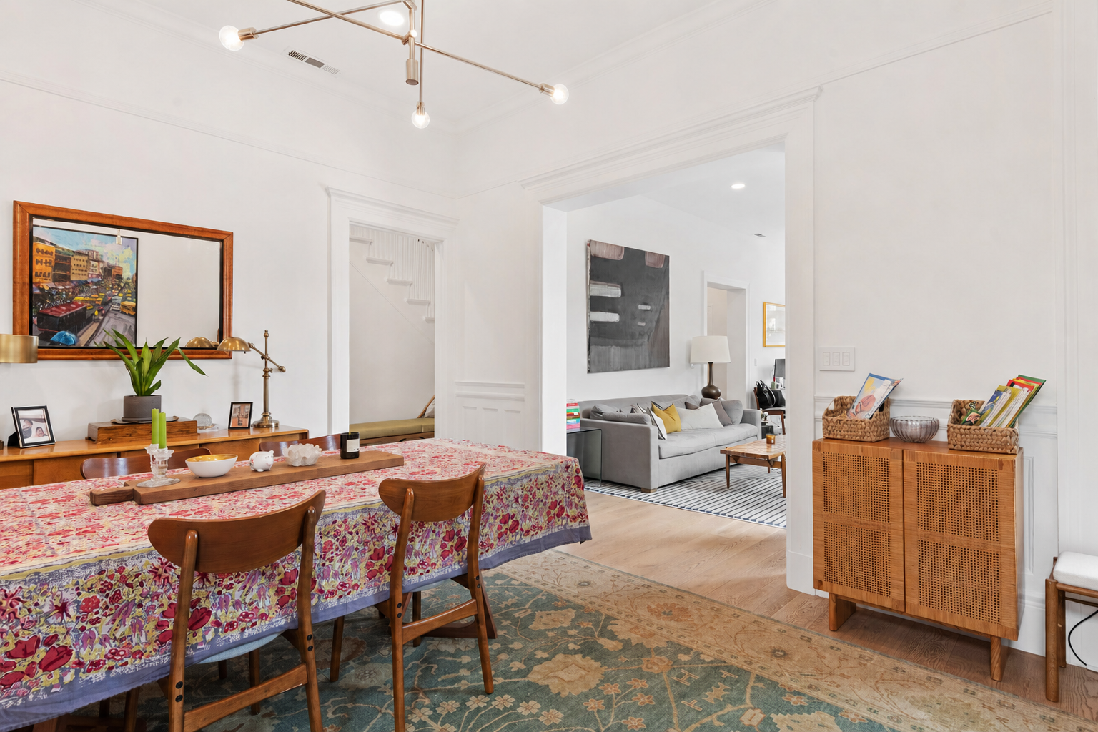 A dining room with a wooden table covered with a colorful tablecloth, surrounded by four wooden chairs. A wooden sideboard on the wall displays a framed picture, a plant in a gray pot, and decorative items. In the adjoining living room, a gray sofa w