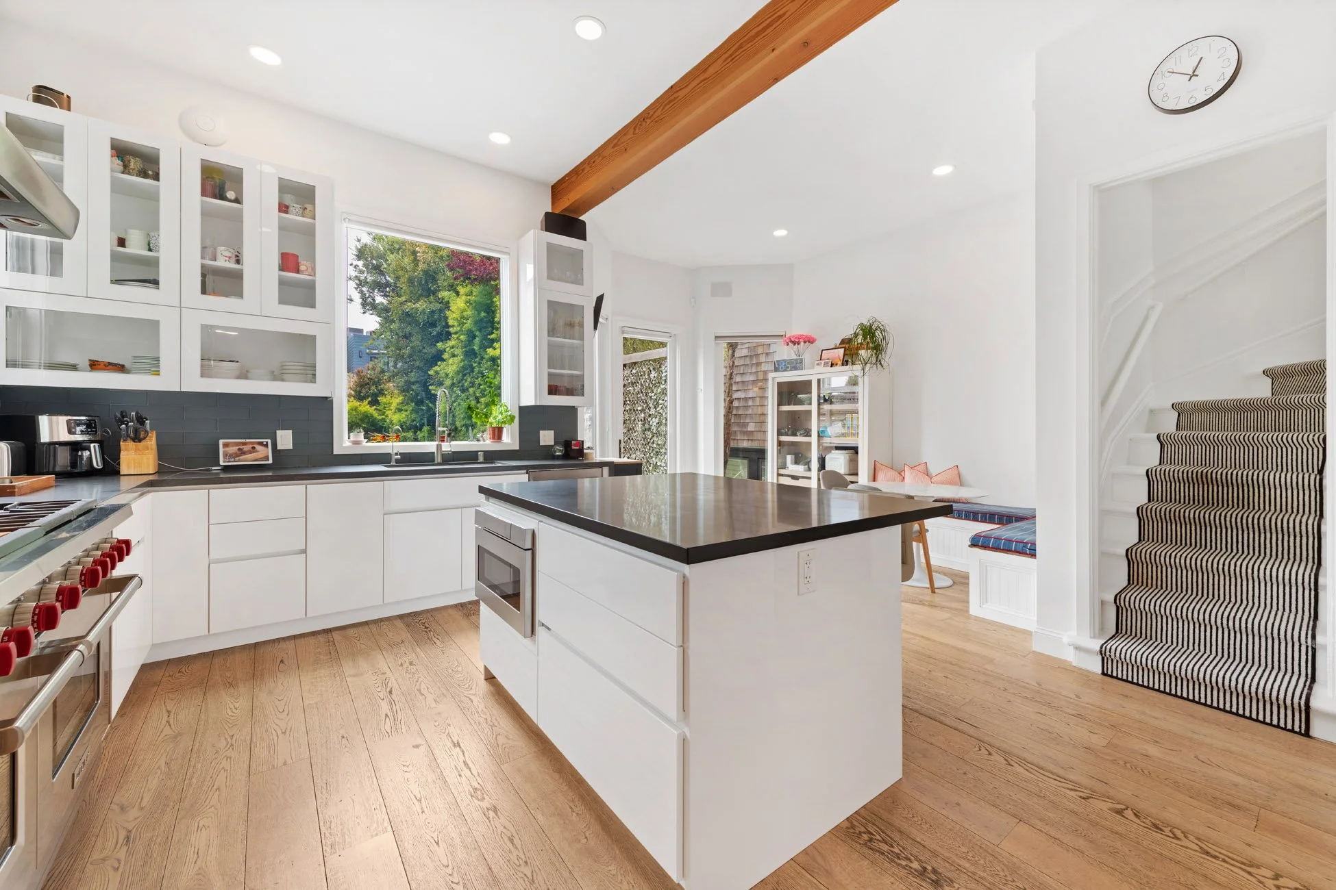 Modern kitchen with white cabinets, black countertops, and a central island with microwave. Large window with outdoor view, wooden floor, staircase with striped carpet, and a seating nook nearby.