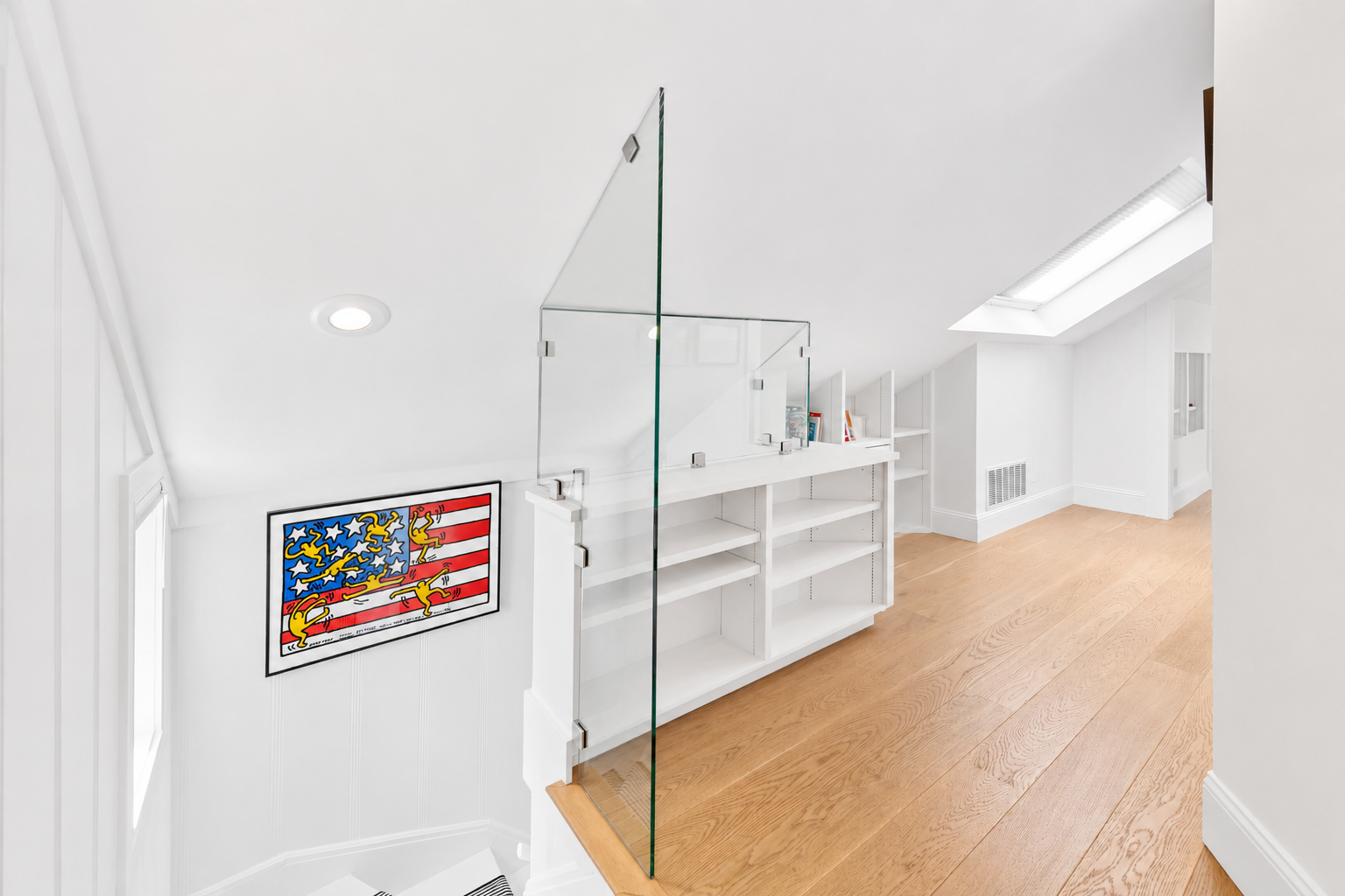 Bright attic space with white walls and built-in white shelves, a skylight window, and a colorful art print of an American flag with yellow creatures. Glass railing separates area from staircase.