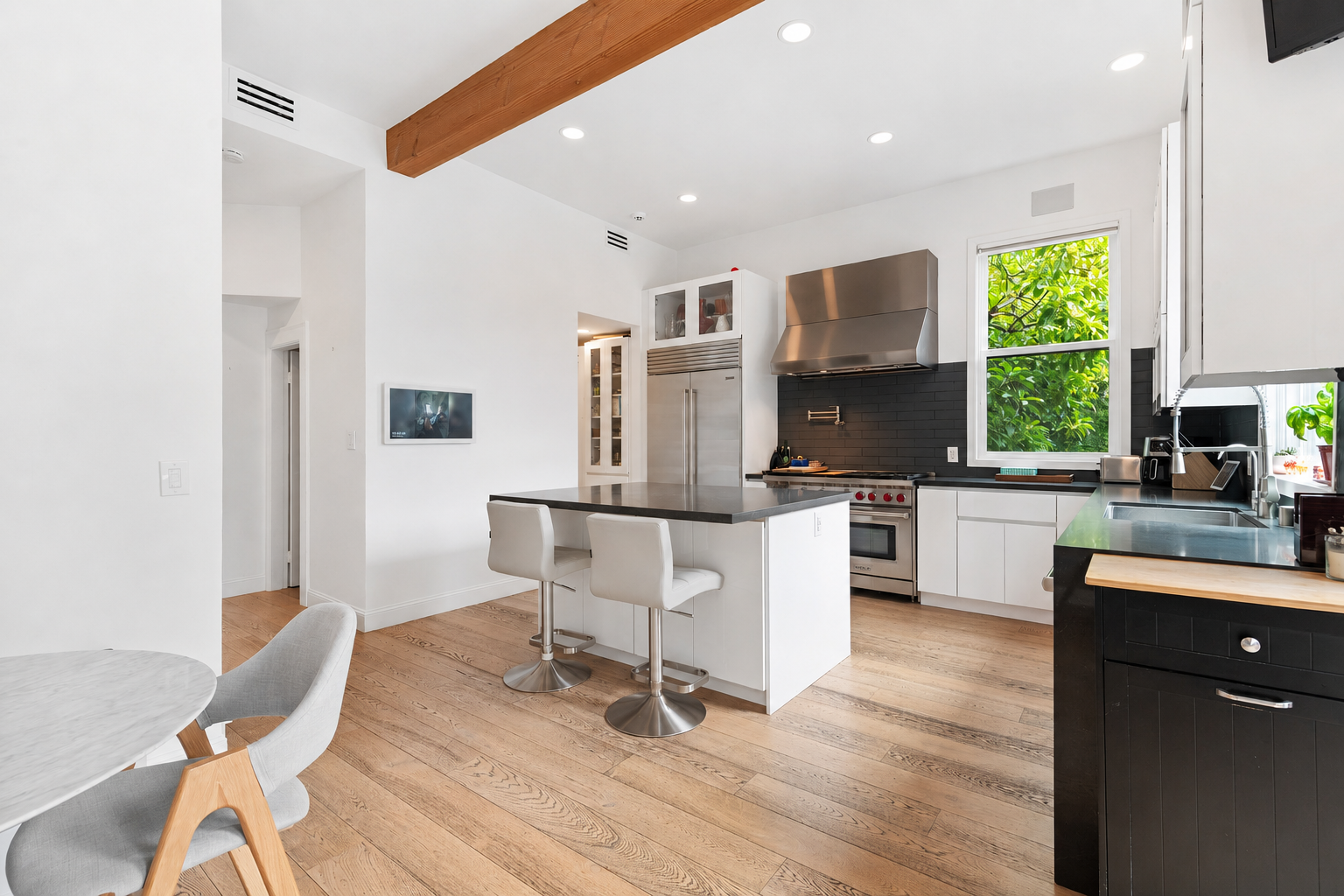 Modern kitchen with white cabinets, black countertops, stainless steel appliances, wood flooring, and a large window showing green foliage outside.