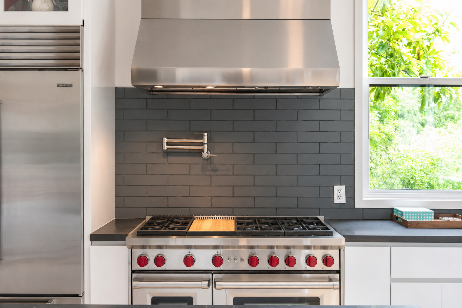 Modern kitchen with black brick backsplash, stainless steel range oven with red knobs, range hood, window with outside greenery, and countertop with storage box.