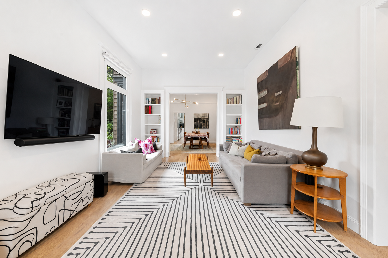 Living room with white walls, large window, wall-mounted TV, white sofa with pink pillows, gray sofa with yellow and neutral pillows, black and white striped rug, wooden coffee table, side table with large lamp, abstract artwork on wall, bookshelf in