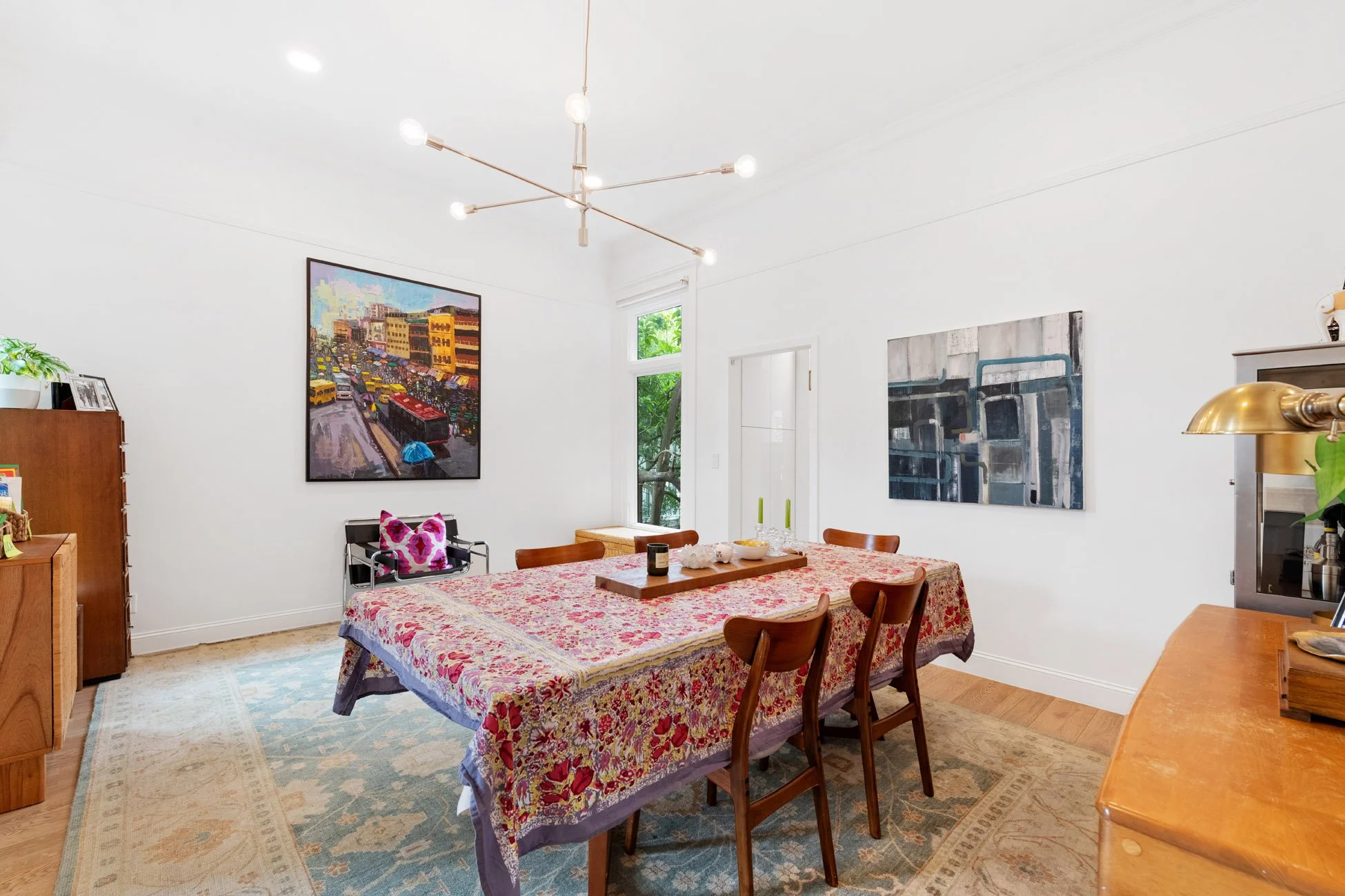A dining room with a rectangular table covered with a floral tablecloth and wooden chairs around it. The wall has two paintings: one depicting city traffic and the other with abstract industrial pipes. A window with greenery outside is in the corner,