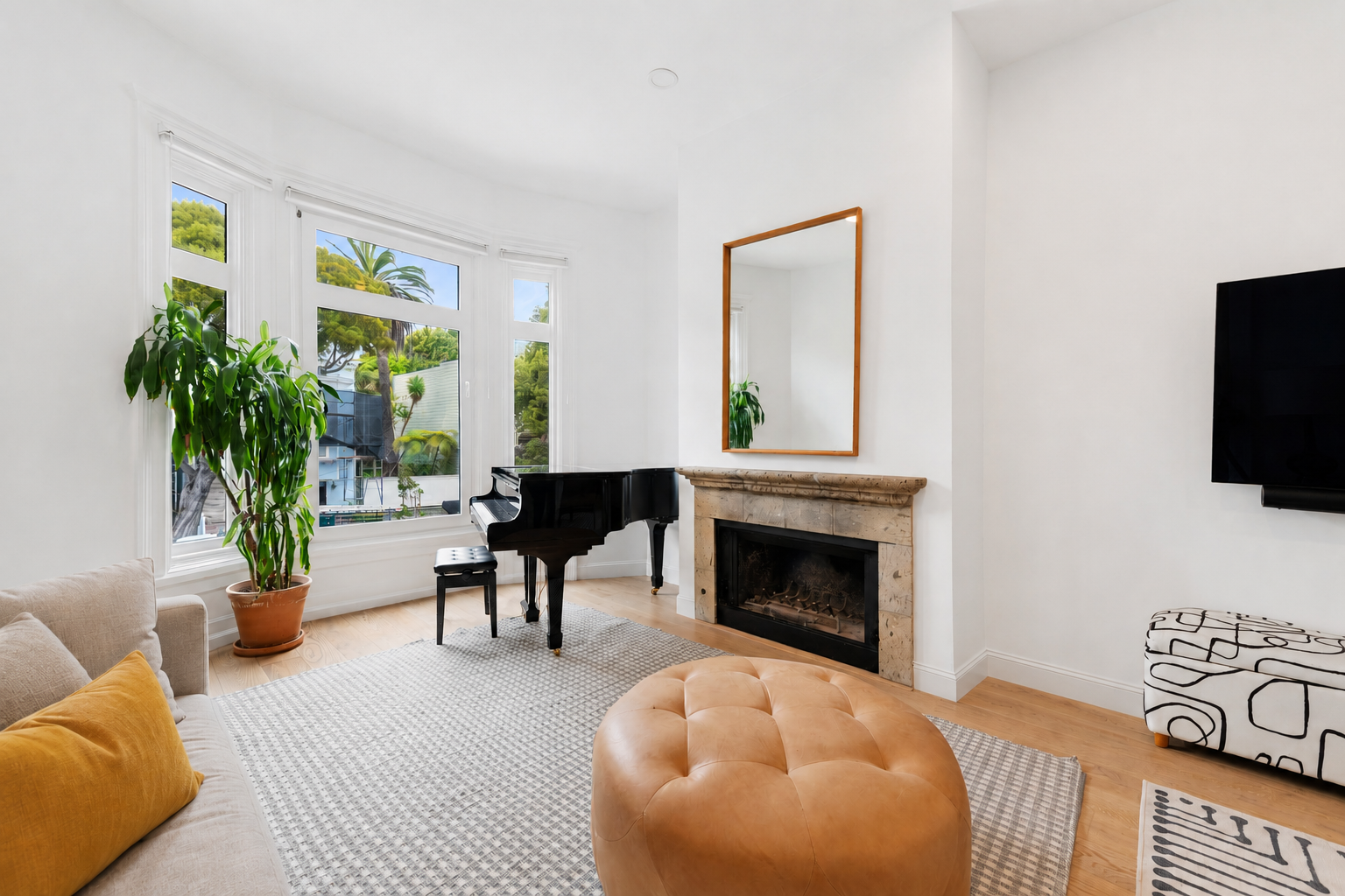 Living room with large window, potted plant, black piano, fireplace, mirror, wall-mounted TV, beige sofa, yellow pillow, patterned ottoman, and white walls.