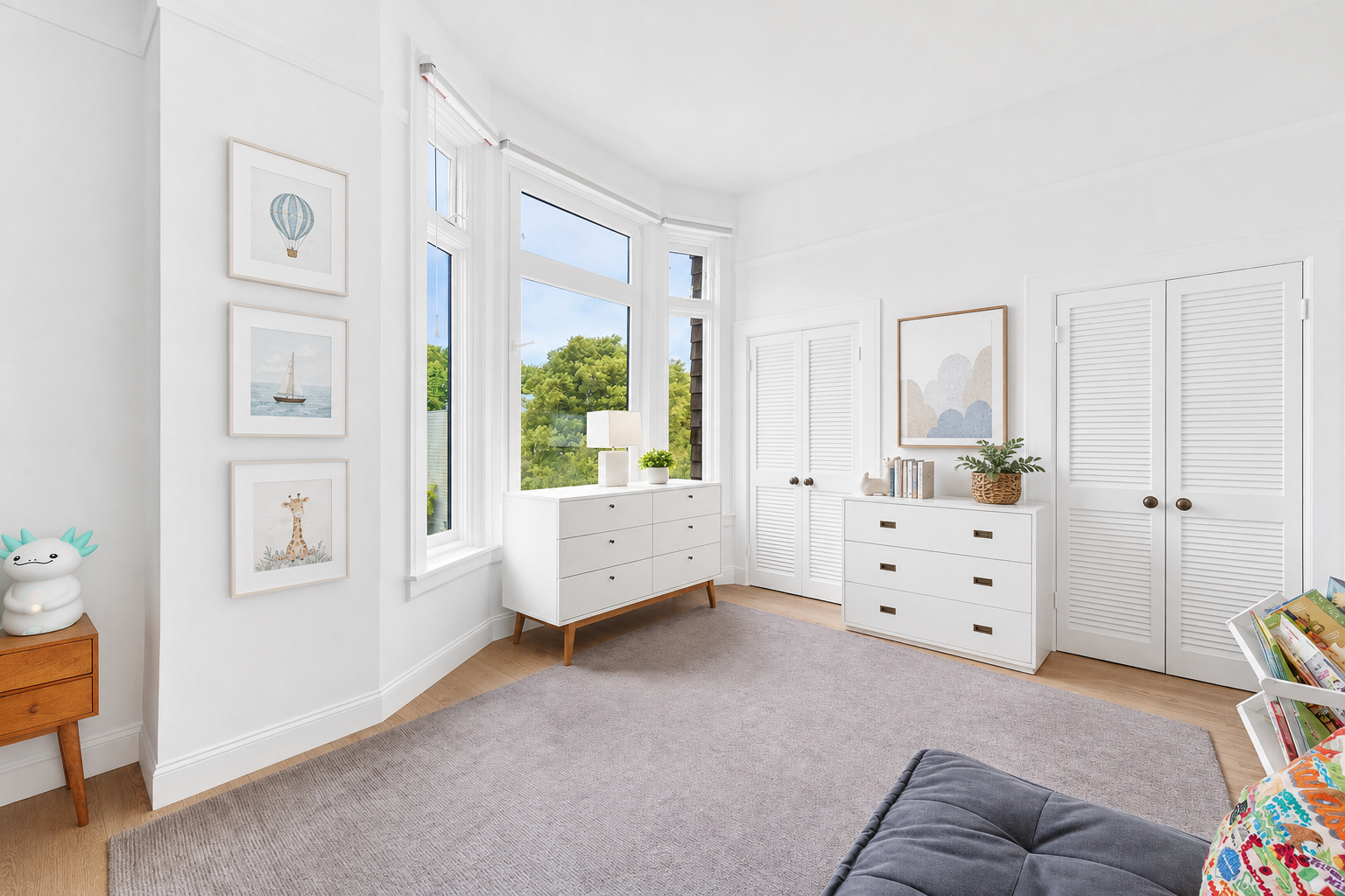 Bright living room with white walls, large windows showing green trees outside, white dresser, bookshelf, and a gray couch with colorful pillows.