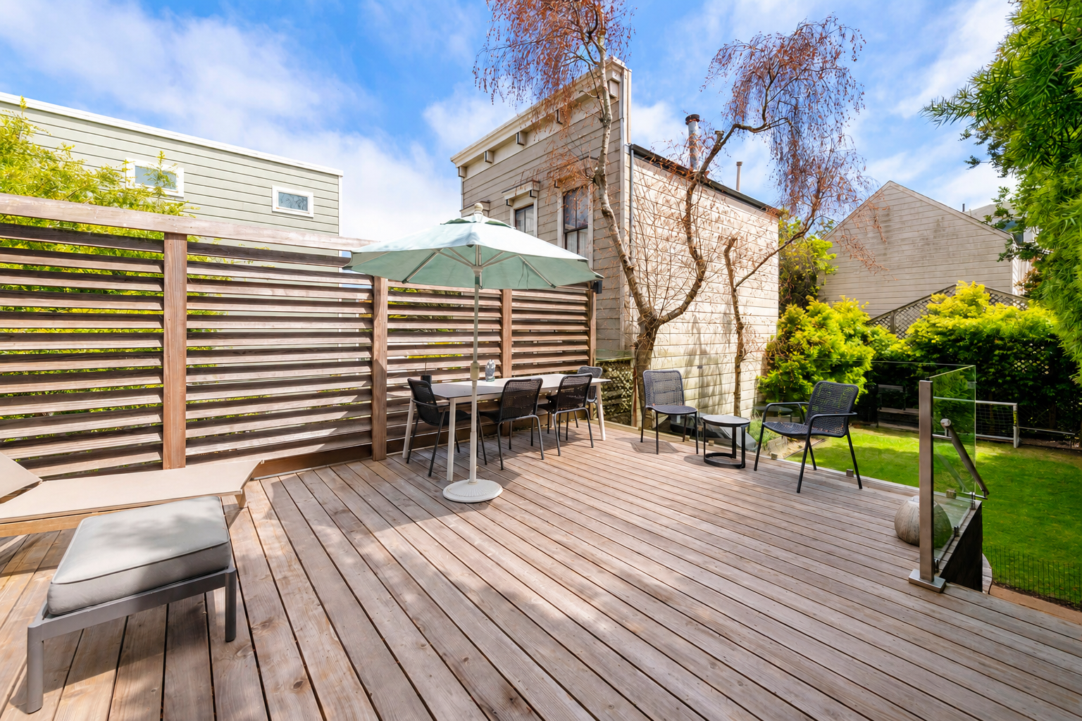 Backyard patio with wooden deck, outdoor furniture, umbrella, chair, and surrounding trees and bushes under a partly cloudy sky.