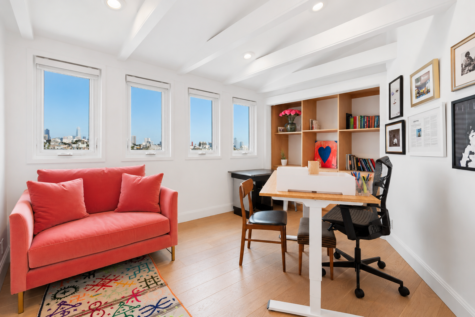 Bright home office with a coral loveseat, wooden floor, white walls, a set of four windows showing a city skyline, a patterned colorful rug, a wooden desk with office supplies, and a wall decorated with framed artwork.