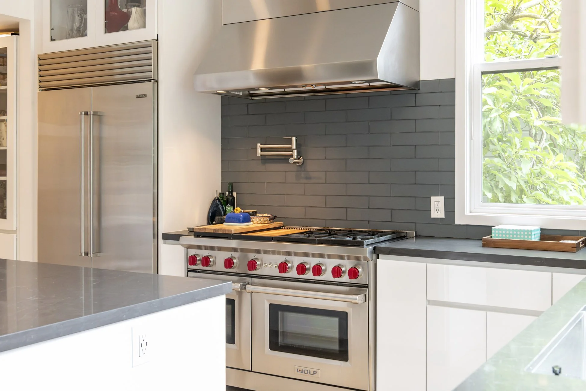Modern kitchen with stainless steel refrigerator, black backsplash, gas stove with red knobs, and a window with greenery outside.