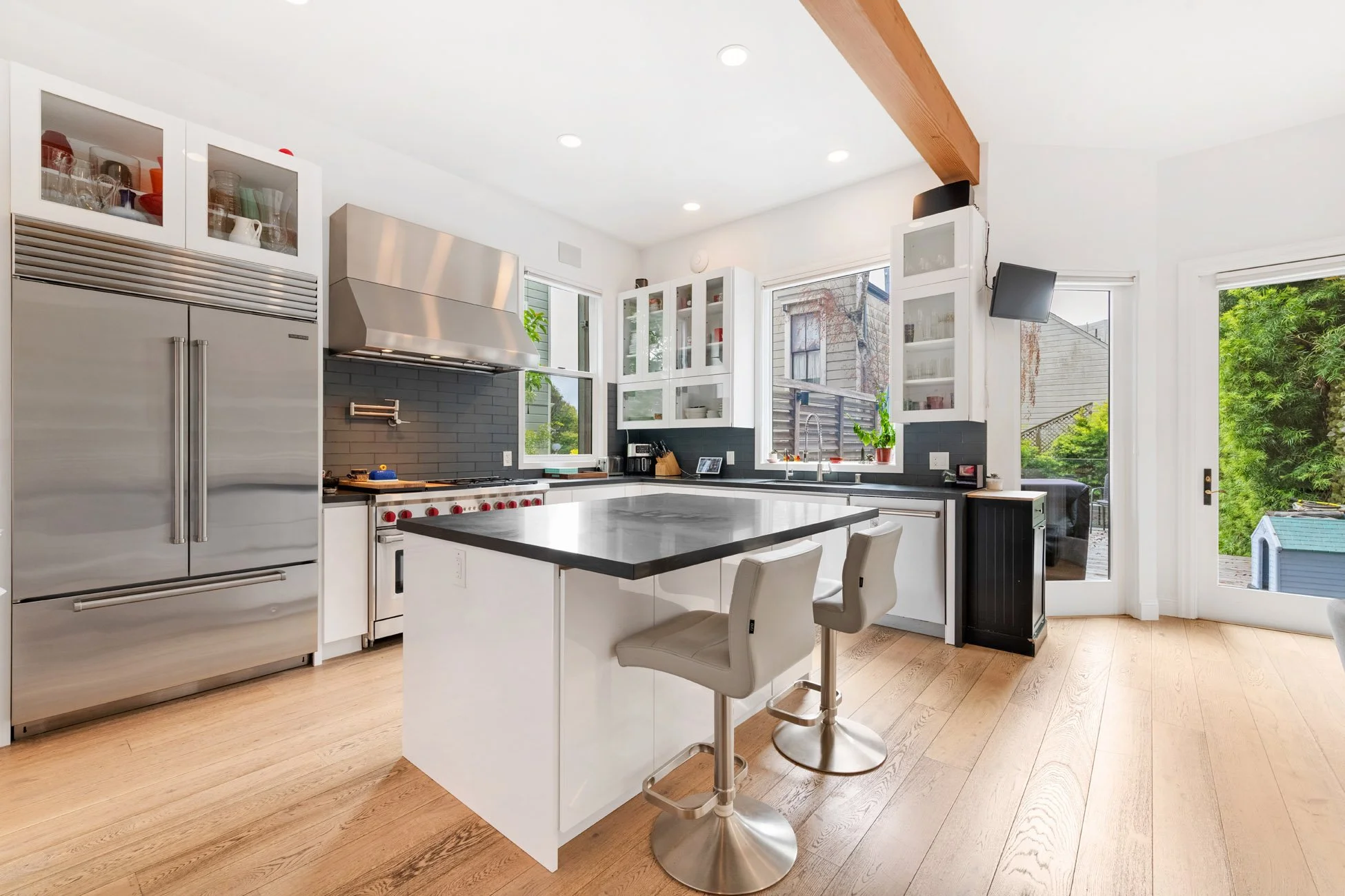 Modern kitchen with white cabinets, stainless steel appliances, a black countertop island, and light wooden flooring, with large windows and a door opening to a backyard patio.