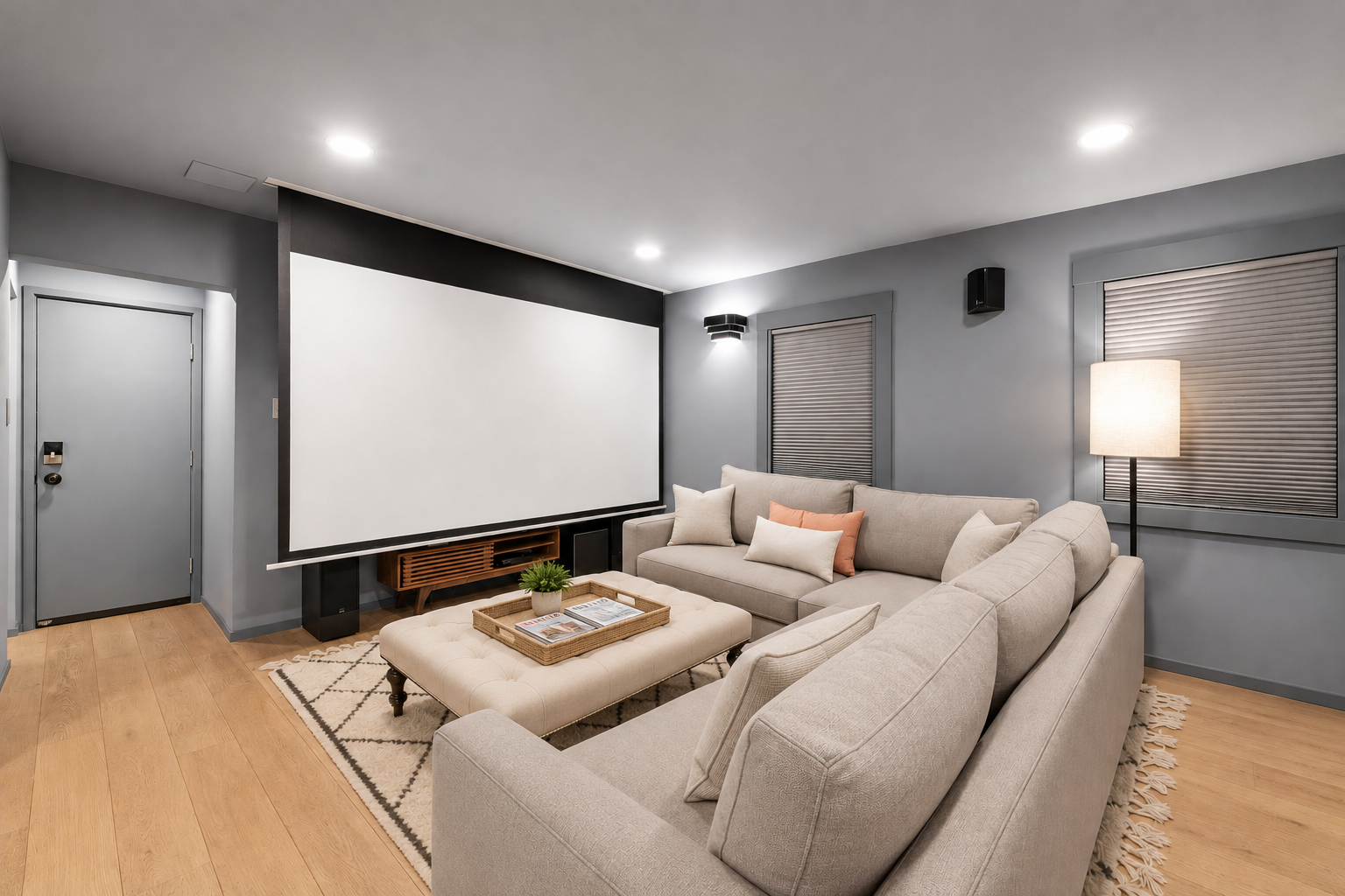 Living room with gray walls, a large beige sectional sofa, a white ottoman with a tray and magazines, a small green plant, and a large white projection screen on the wall.