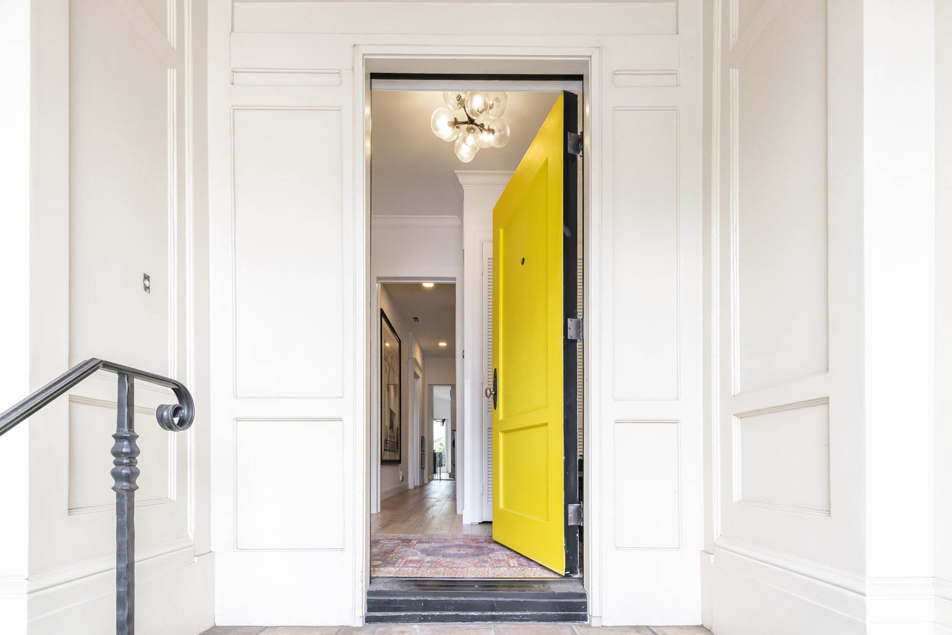 Open yellow front door of a house, view into a hallway with a chandelier and artwork, white walls with decorative panels, and hardwood floors.