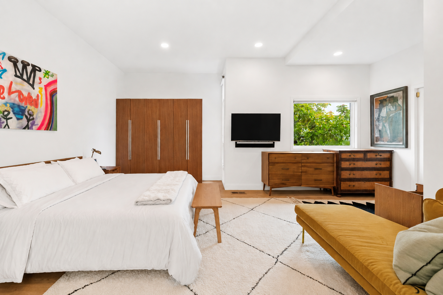 Bright modern bedroom with white walls, wooden furniture, a yellow velvet daybed, a white rug, and large window with greenery outside.