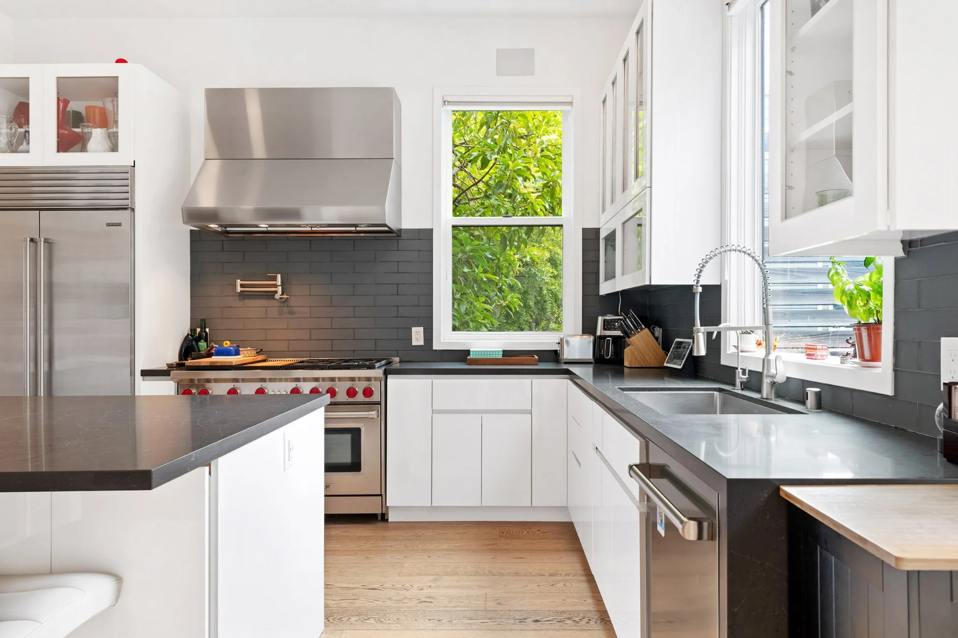 Modern kitchen with white cabinets, black countertops, stainless steel appliances, and a large window showing green foliage outside.