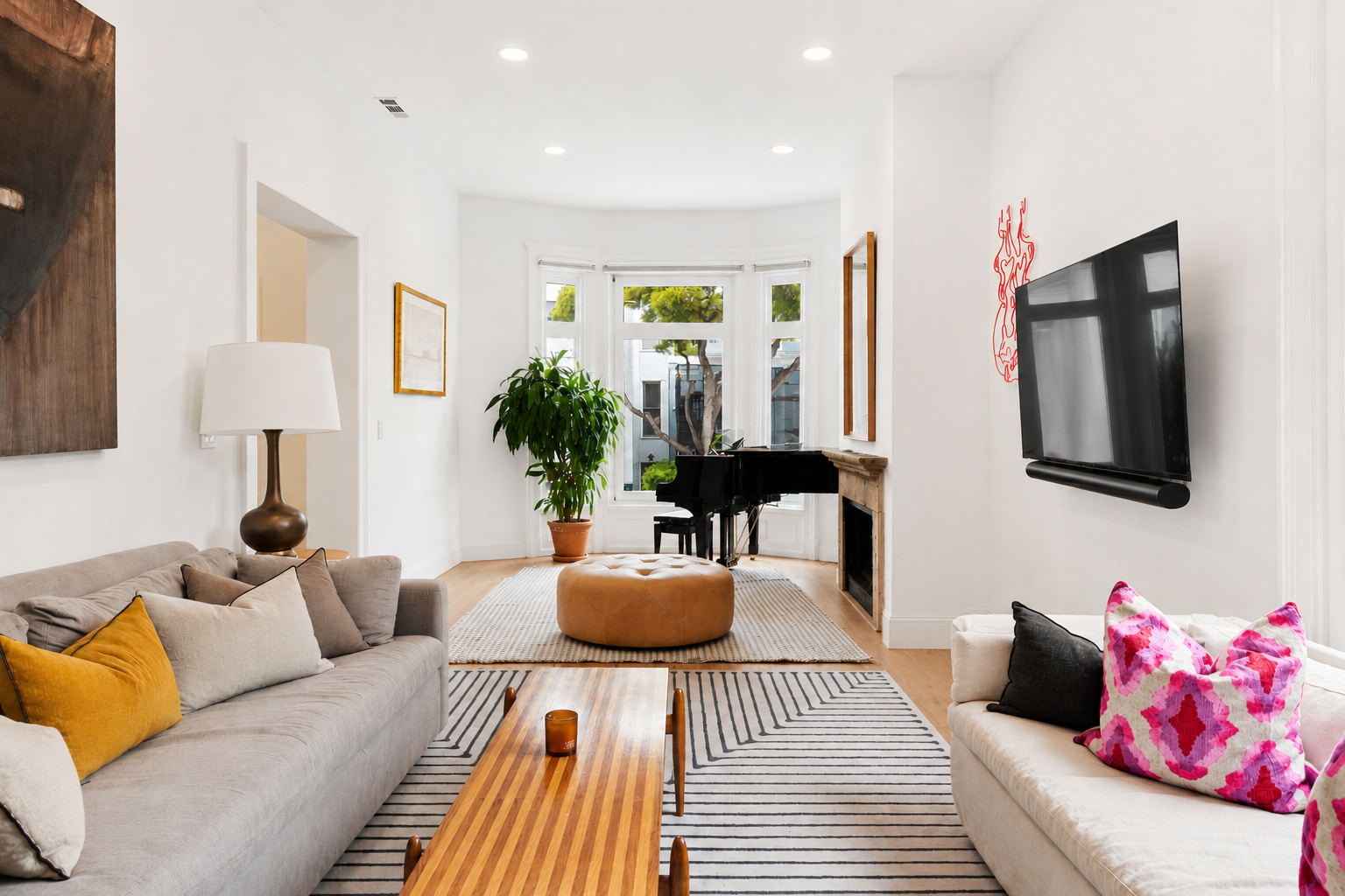 Bright living room with two sofas, a coffee table, a grand piano near the large bay window, a potted plant, a fireplace, a wall-mounted TV, and various colorful pillows and decor.
