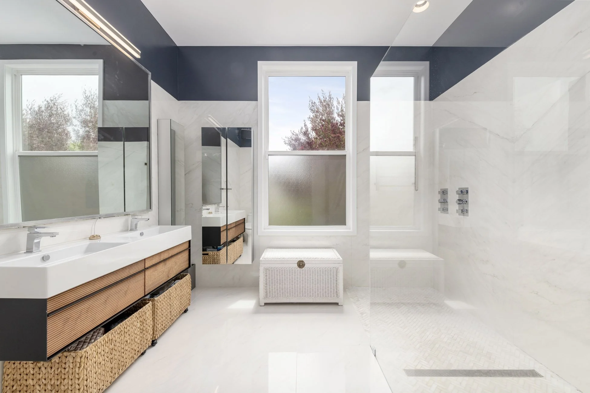 Modern bathroom with white and dark gray walls, double sinks, wicker storage baskets, large windows with views of trees, and a walk-in shower with a glass door.