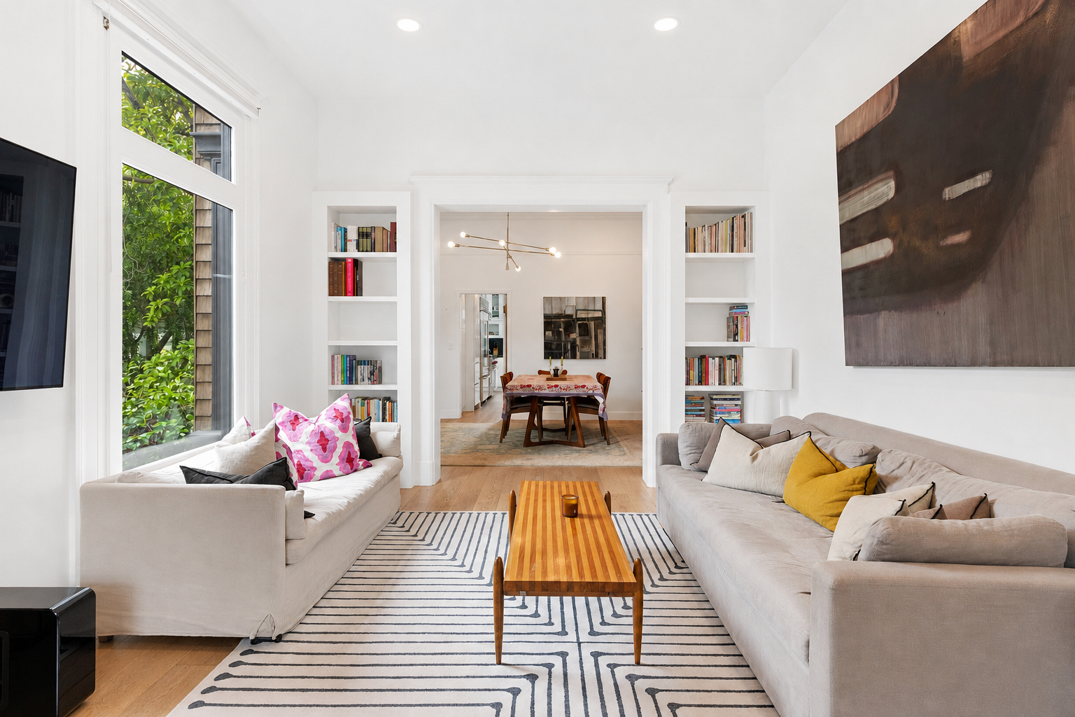 Living room with large window, two cream sofas with colorful cushions, a striped rug, and a wooden coffee table. Bookshelves on each side of an opening lead to a dining room with a table, chairs, and a modern chandelier.
