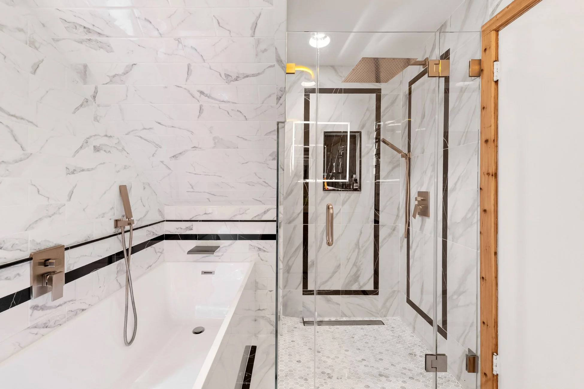 A modern bathroom with white marble walls and a glass shower enclosure with black trim. The shower has a large rainfall showerhead and a handheld showerhead. There is a small window in the shower area, and the entrance has a wooden door frame. The ba