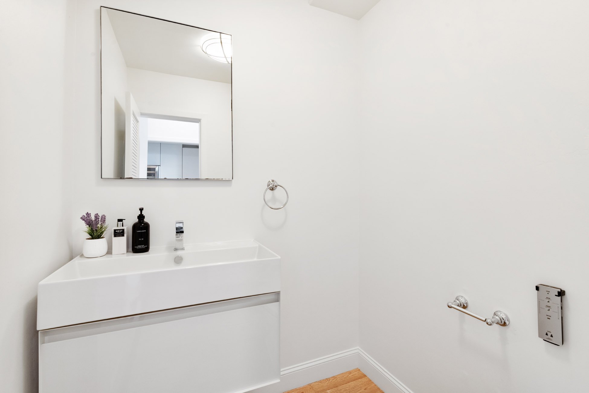 Minimalist bathroom with a white vanity and sink, a rectangular mirror, black soap dispenser, lavender plant, and toiletries, white walls, chrome towel rack, and a small wall fixture.
