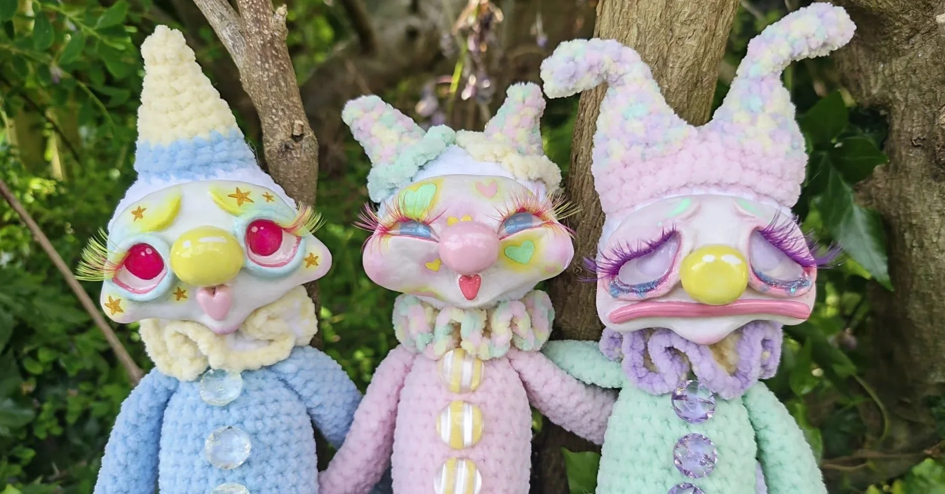 Three whimsical plush dolls with masks resembling clowns or fantasy characters are hanging on a tree branch. They wear pastel-colored fuzzy hats with bunny ear shapes and have exaggerated facial features with long false eyelashes, glitter, and large decorative eyes.