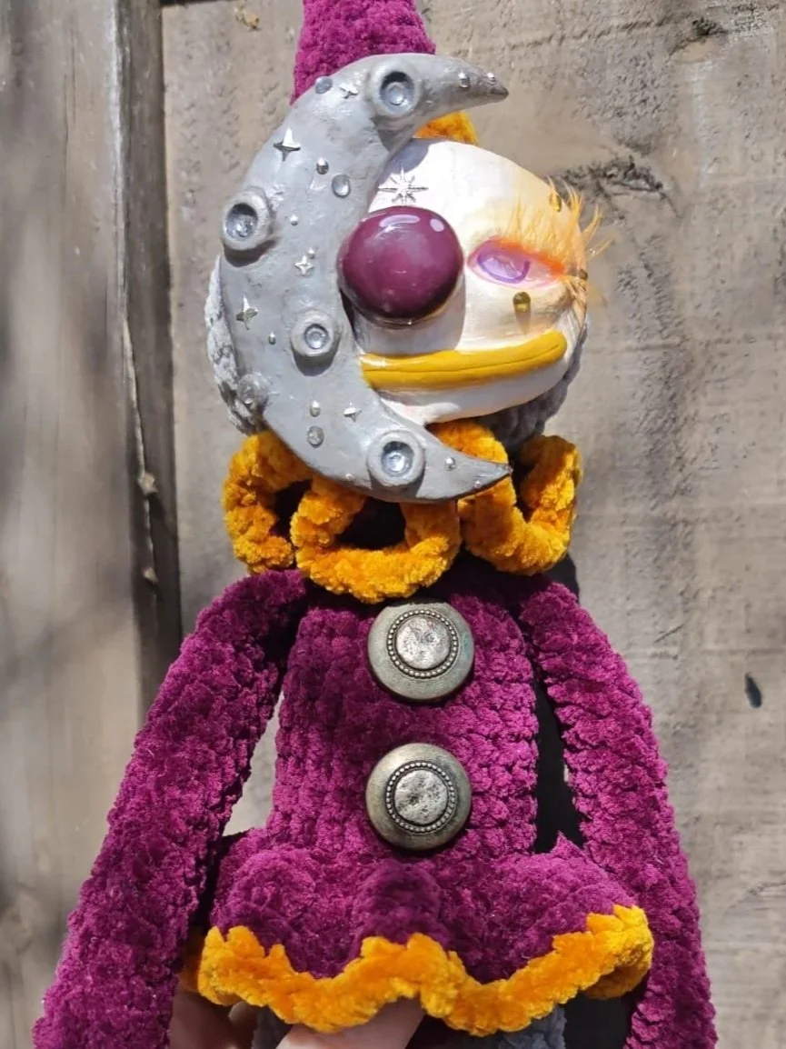 A whimsical handmade doll with a clown face and colorful clothing. The face is painted with exaggerated features, including a purple nose, and the doll has a moon-shaped headpiece with decorative elements. It is dressed in a textured purple fabric with yellow accents and large decorative buttons. The background is a plain wooden wall.