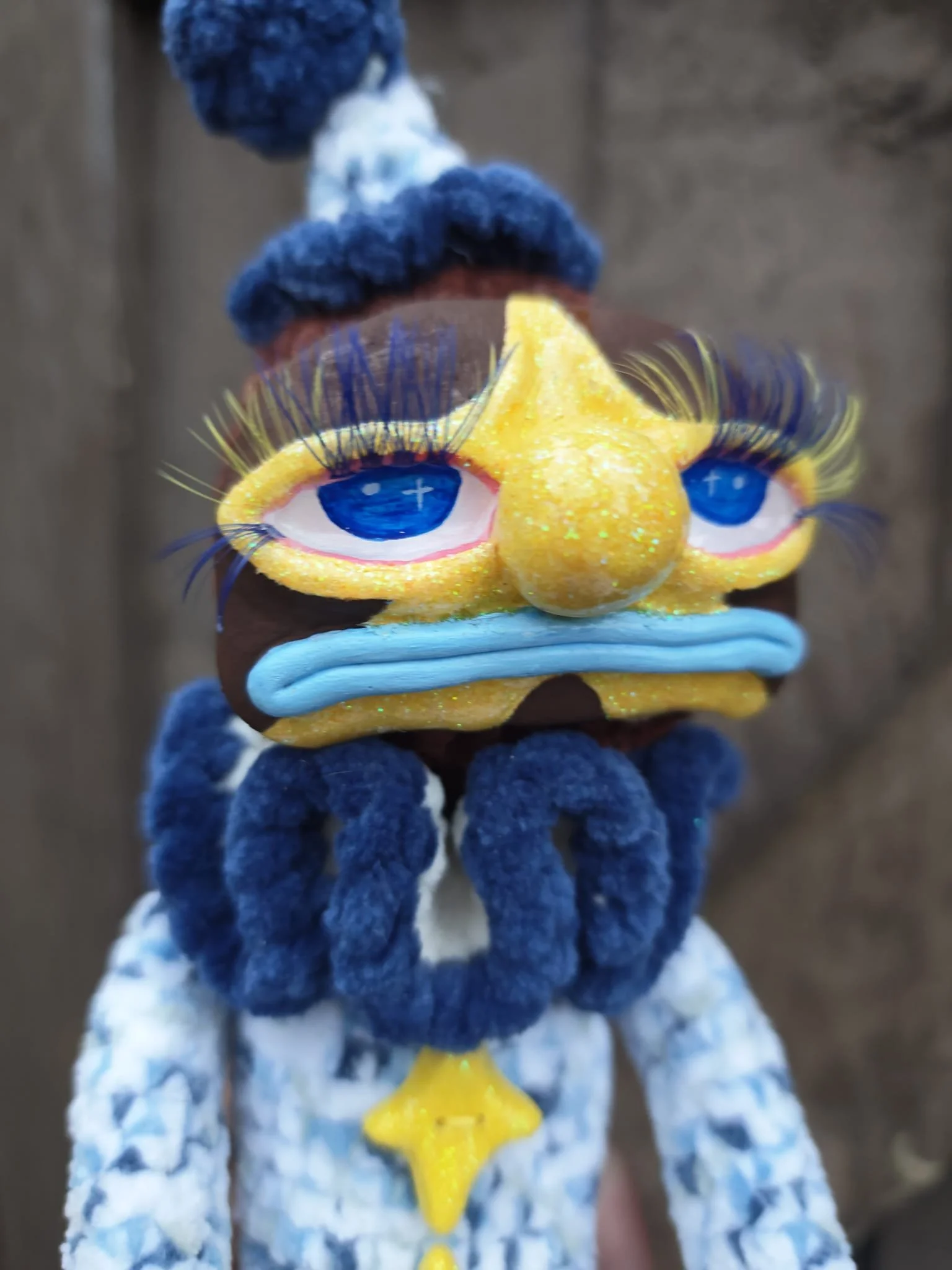 A close-up of a handmade craft resembling a colorful cartoon bird with exaggerated blue eyes, yellow beak, and fluffy blue hair, held by a person dressed in a blue and white patterned outfit.