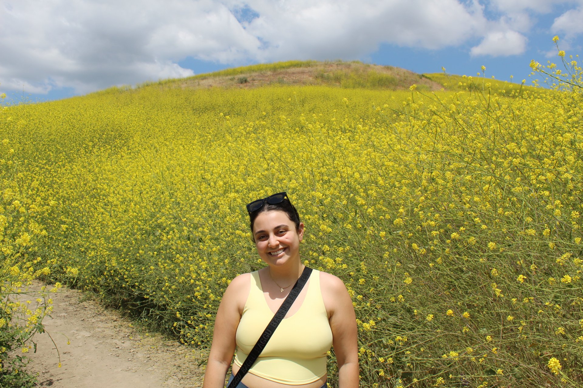 Young woman smiling in a yellow top, standing in front of a large field of yellow flowers with a hill in the background under a blue sky with clouds.