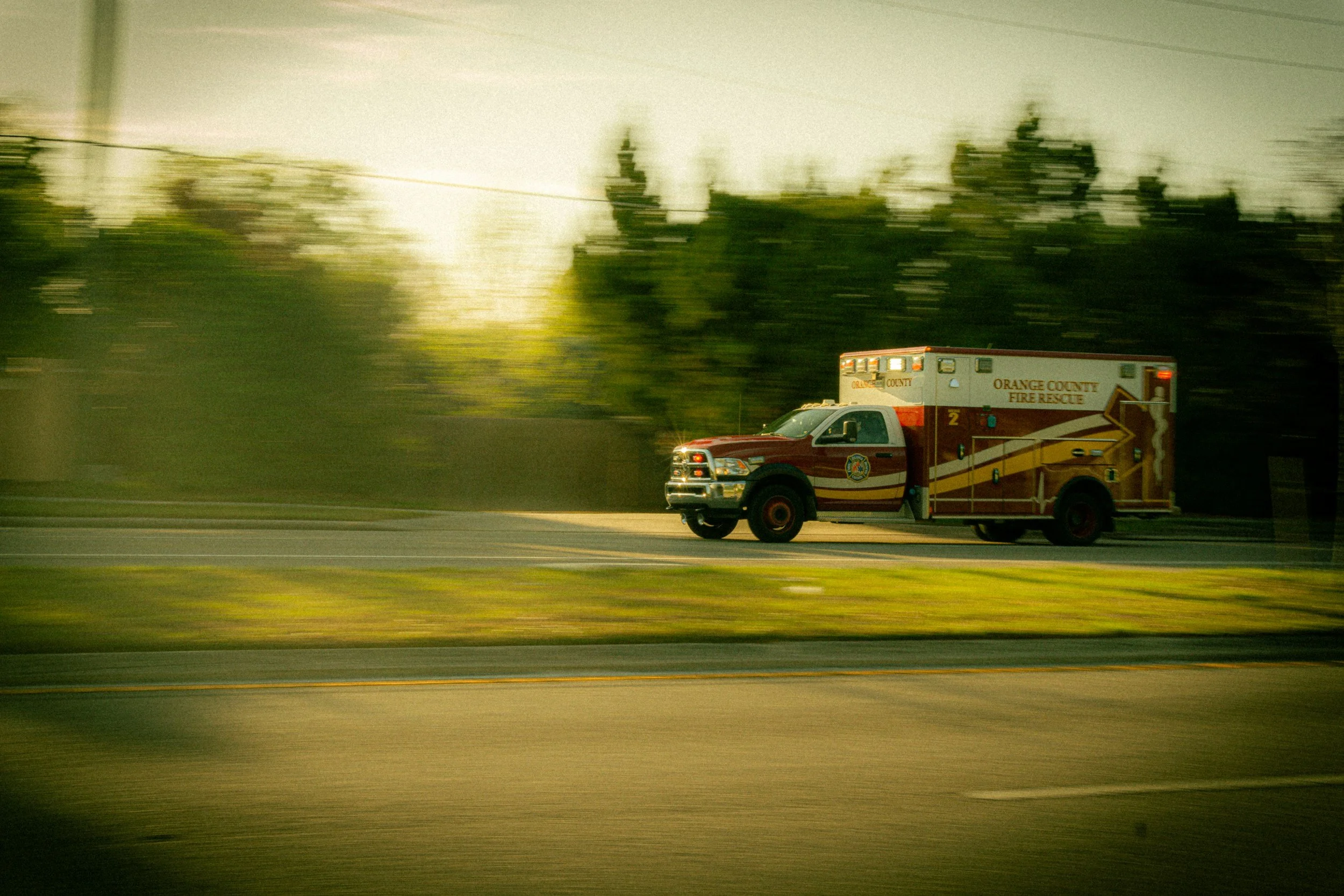 A moving orange county fire rescue vehicle on a highway, with trees and power lines in the background and the sun shining brightly.