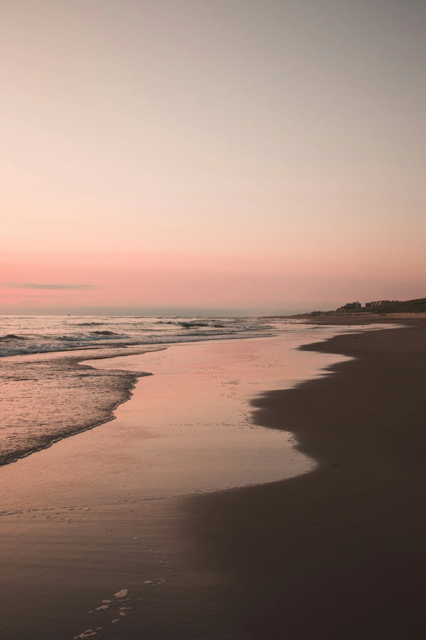 Beach at sunset with gentle waves and footprints in the sand, with buildings visible in the distance.