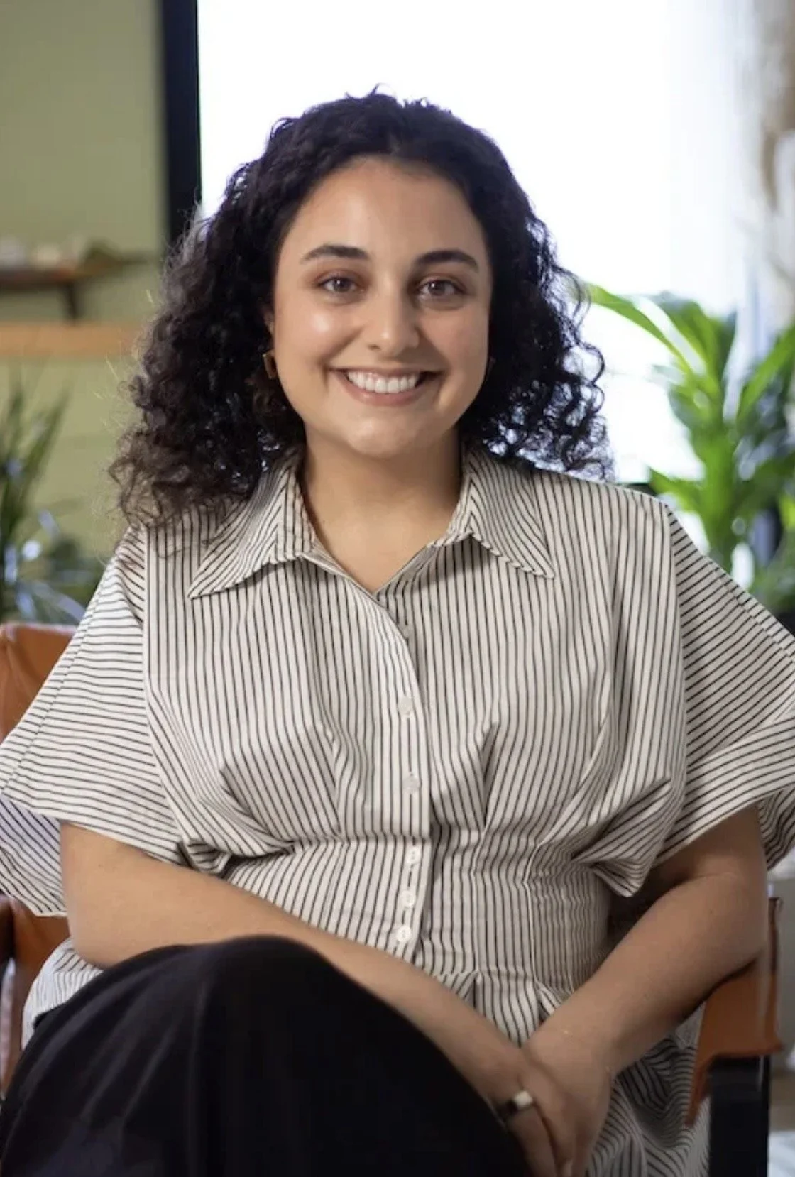 A woman with curly dark hair smiling, wearing a striped button-up shirt, sitting indoors with a background of plants and a TV.