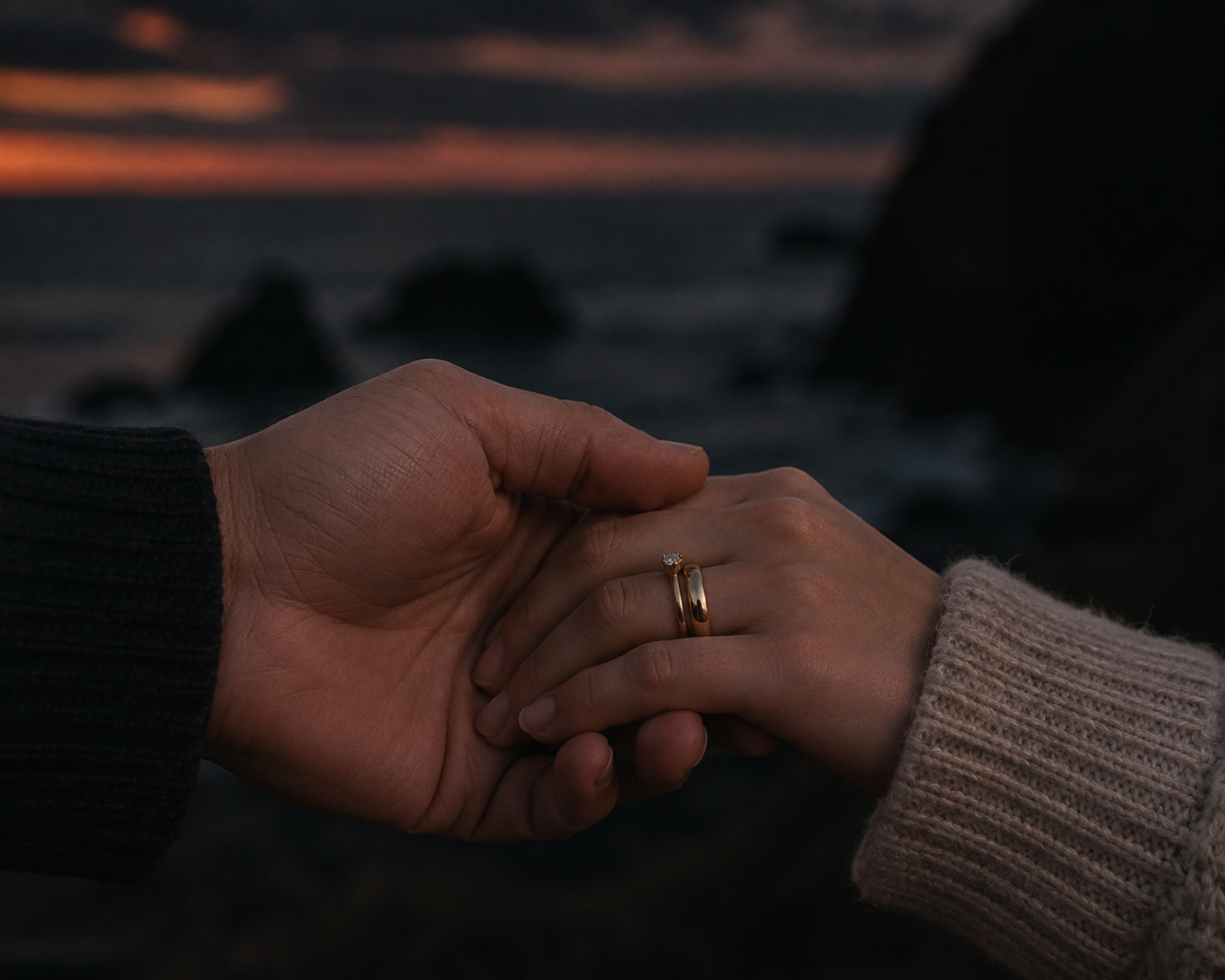 Close-up of a couple holding hands, with a sunset over water in the background. The woman is wearing a beige sweater and an engagement ring, and the man is wearing a black sweater.