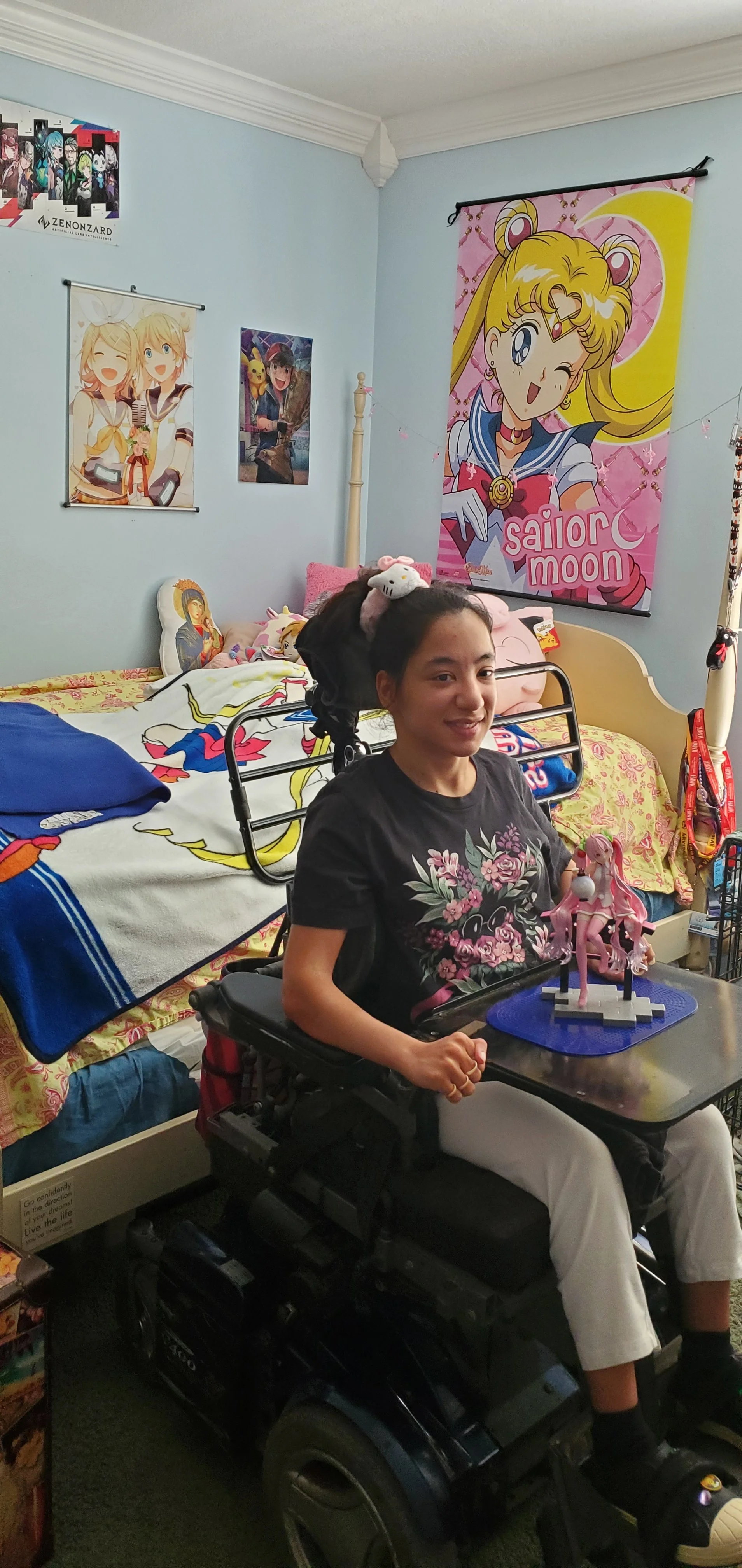 A young woman sitting in a wheelchair in her bedroom, smiling at the camera. The room is decorated with anime posters and character-themed bedding, featuring Sailor Moon and other animated characters. She has a Hello Kitty headband and a floral shirt, and there is a pink anime figurine on a small table in front of her.