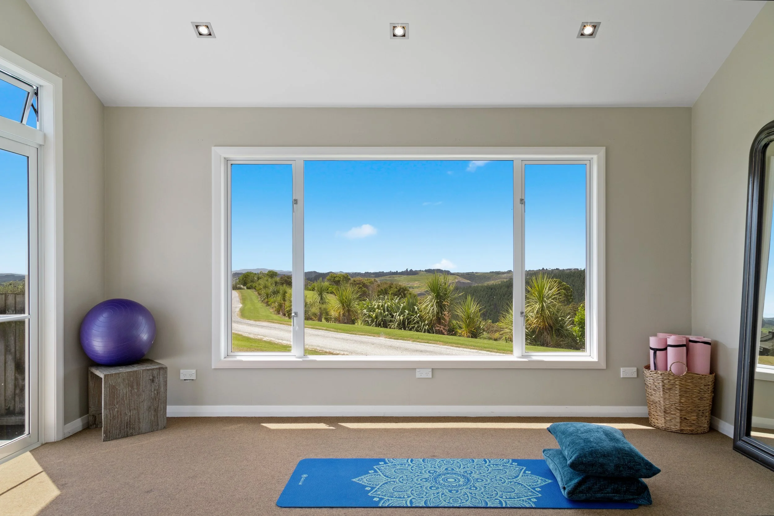 Indoor yoga or exercise room with large window overlooking landscape, purple exercise ball, blue yoga mat, black pillow, pink foam rollers, wicker basket, and mirror.
