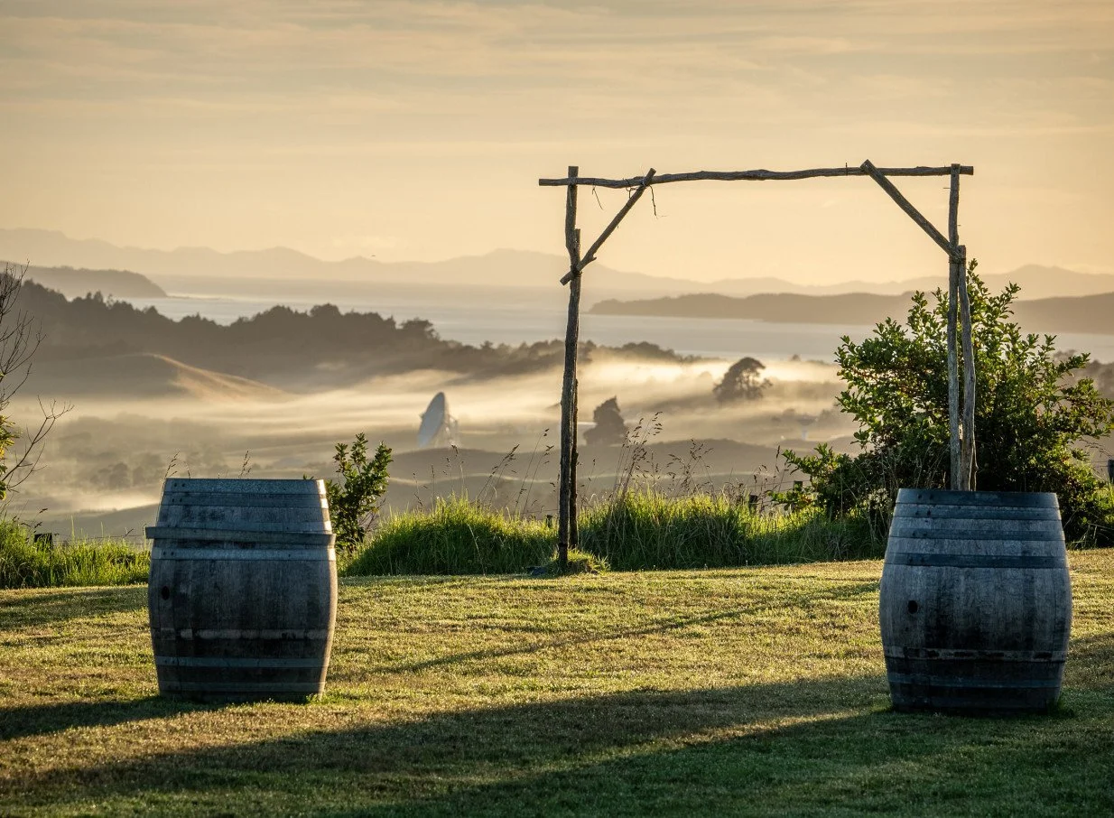 Rustic outdoor ceremony setting framed by wine barrels and timber arch overlooking rolling hills and morning mist
