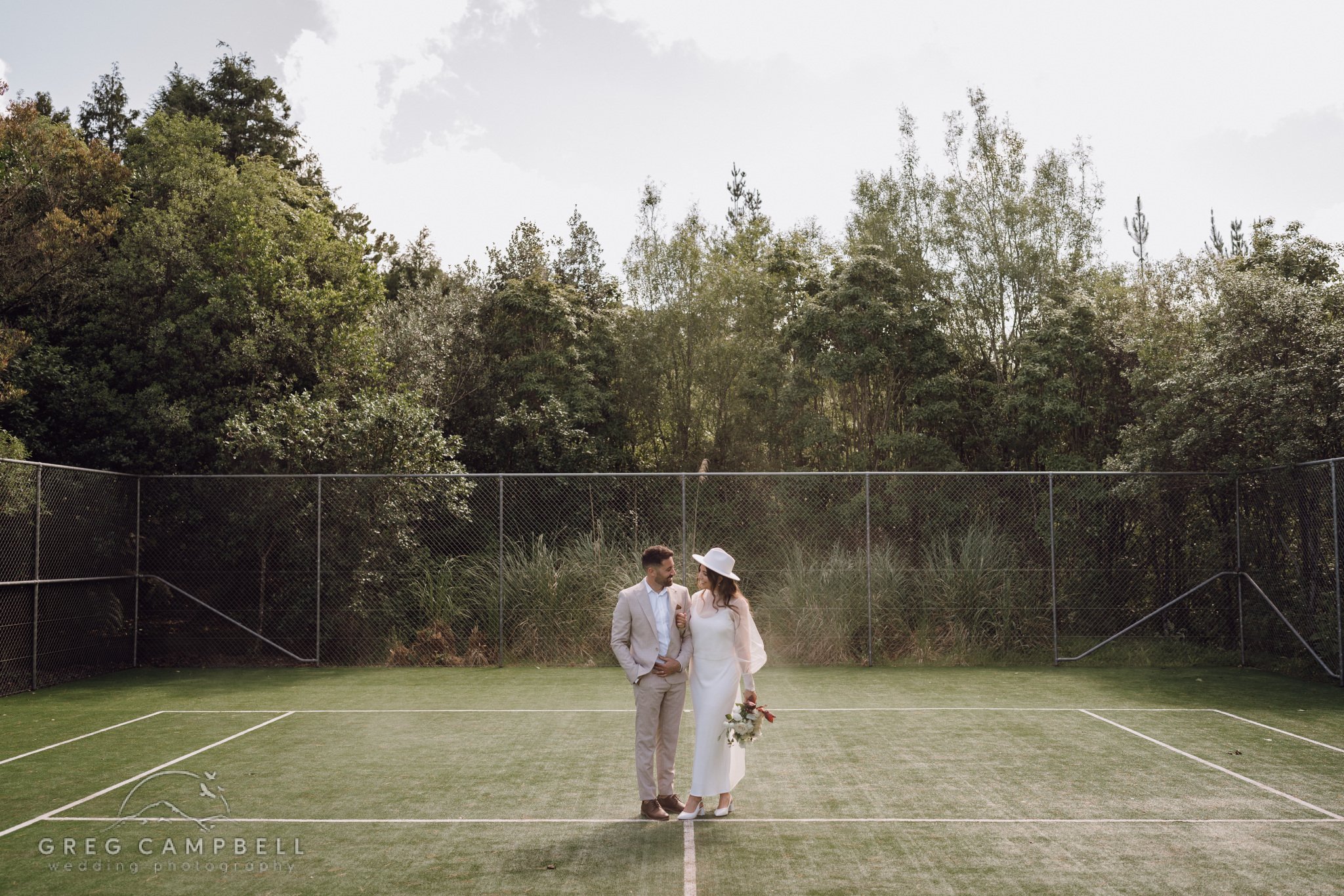 Bride and groom standing together on a grass tennis court, surrounded by trees and countryside.