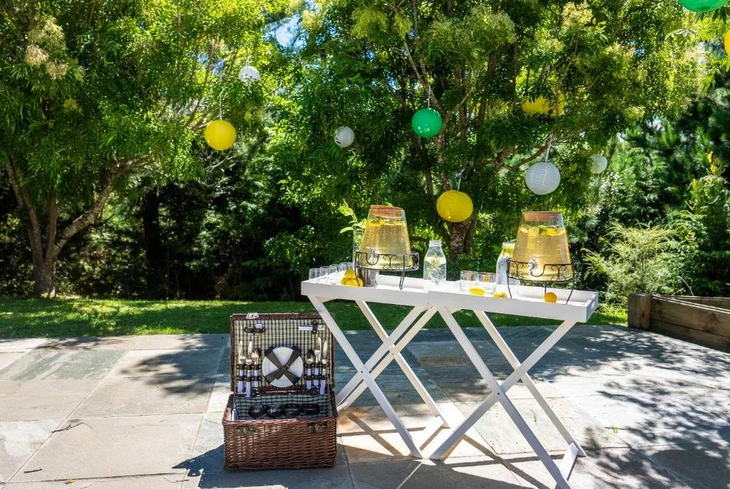 Outdoor garden drinks station with glass beverage dispensers, lemons, picnic basket and hanging lanterns set beneath leafy trees on a sunny day.