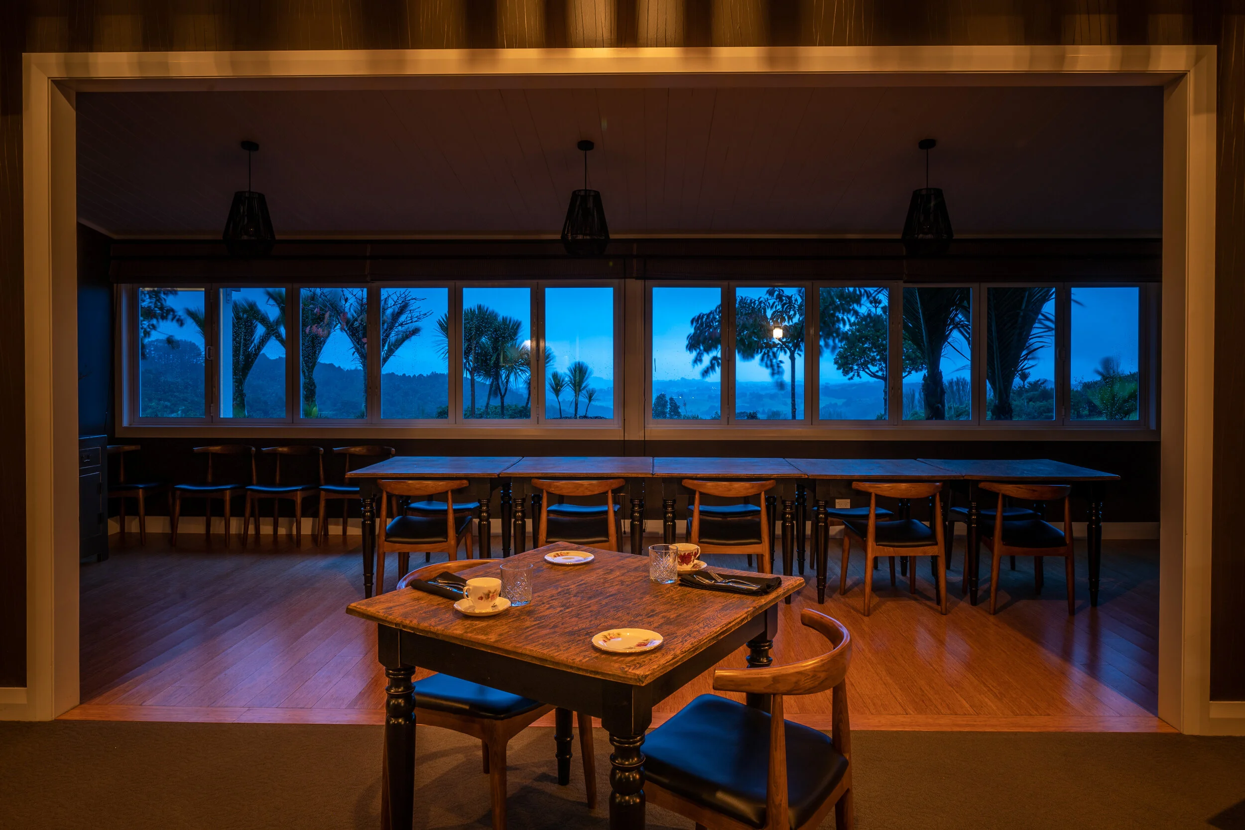 Elegant private dining room with timber tables and chairs, set for dinner with blue-hour views through large windows.