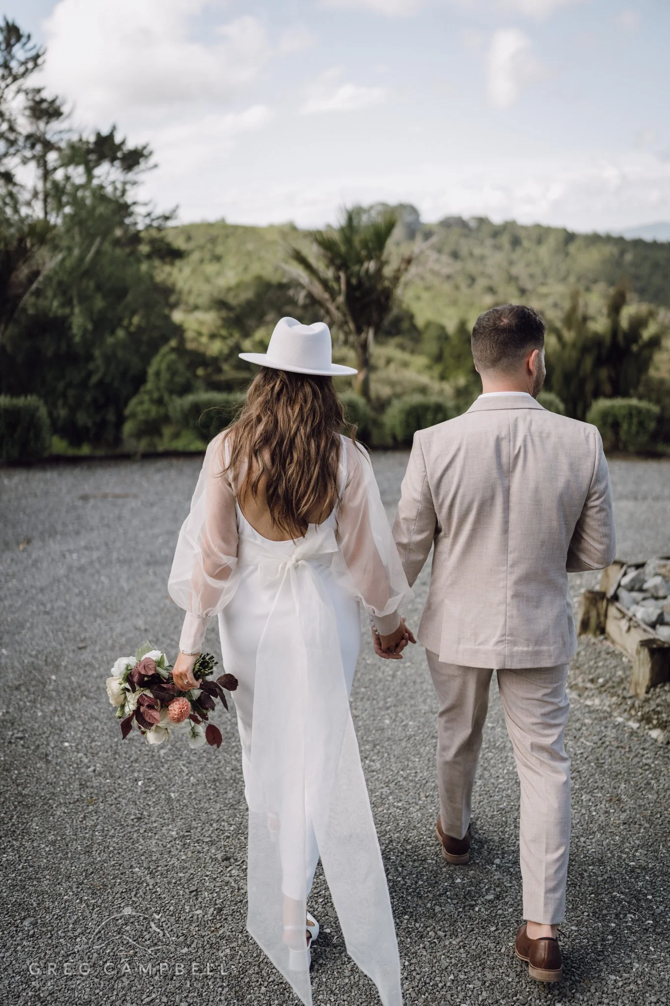 Newlyweds walking hand in hand through a garden path, surrounded by nature and soft afternoon light.