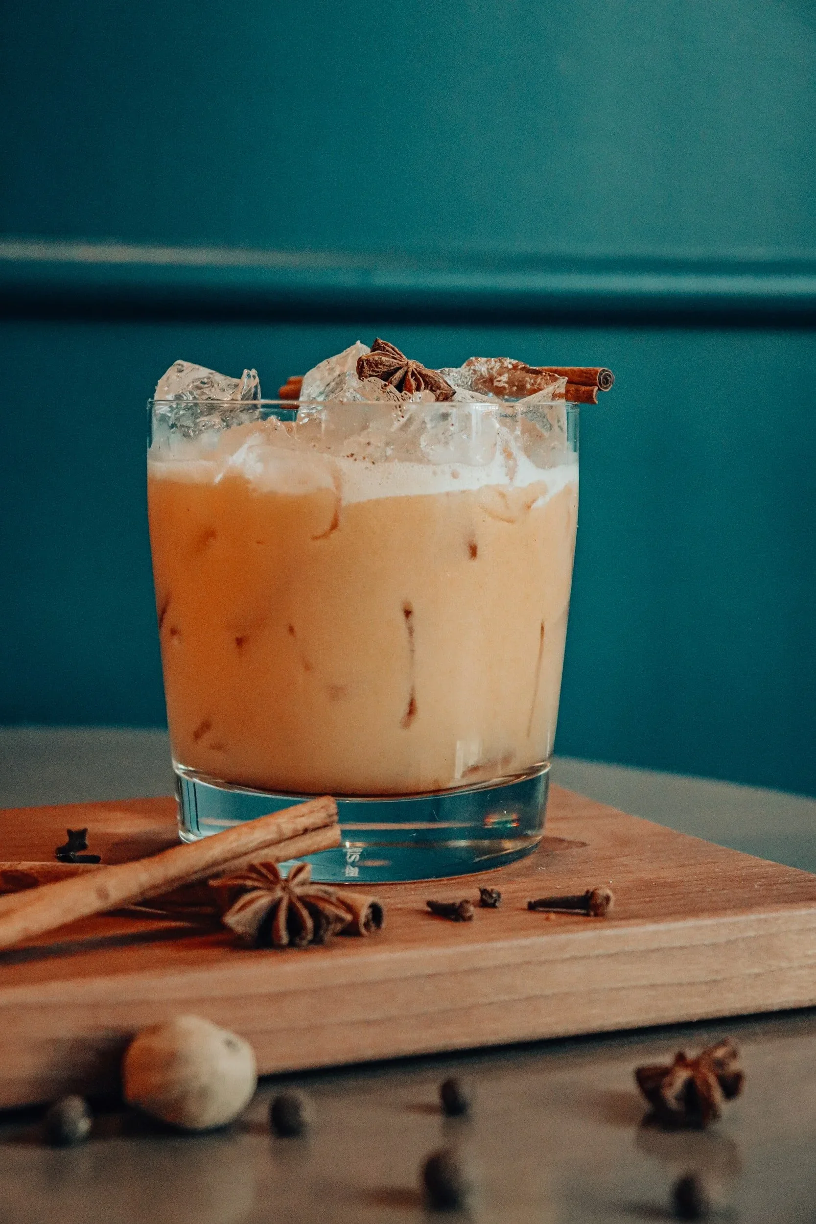 A glass of iced chai tea topped with cinnamon sticks and star anise, with cinnamon sticks and star anise spilled on a wooden surface.