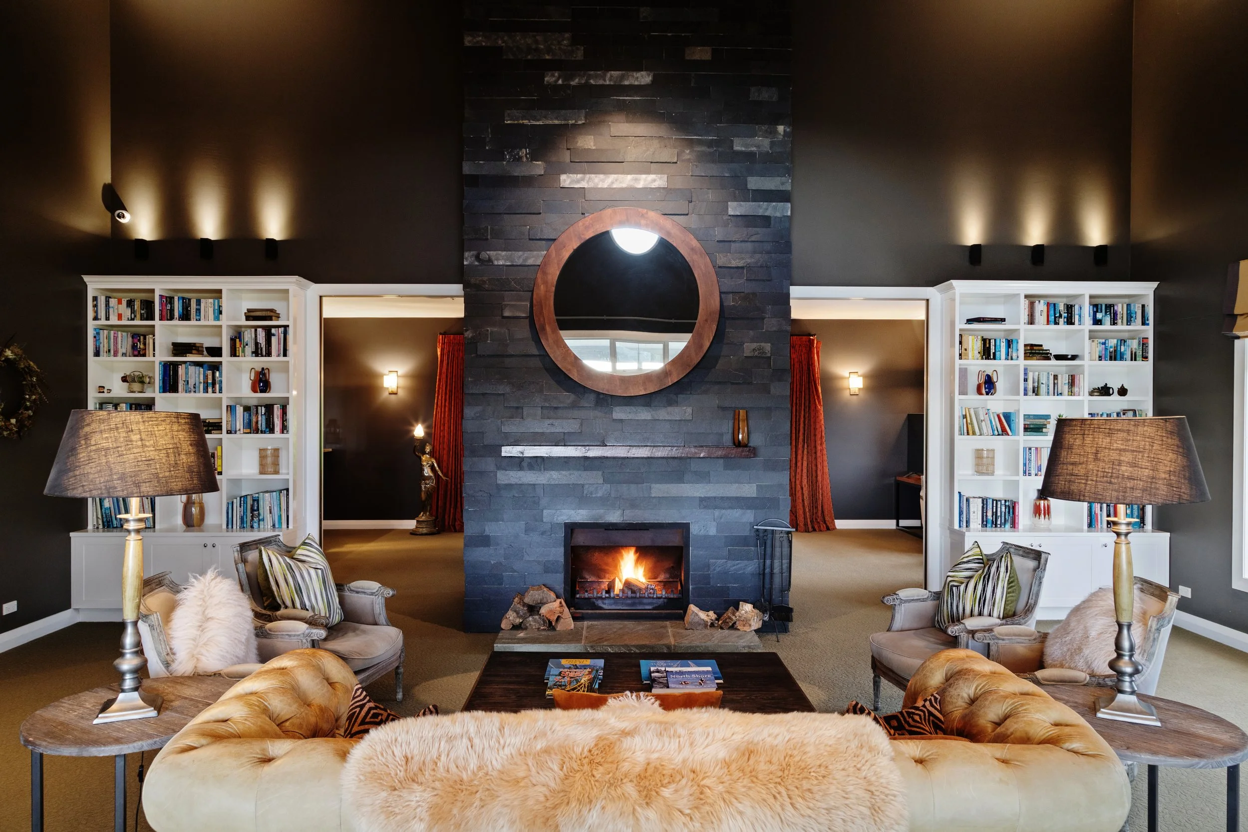 Living room with dark gray stone fireplace, two white bookshelves, two armchairs with striped and velvet cushions, beige tufted sofa with furry throw, round wooden side tables, lamps, and a circular mirror above the fireplace.