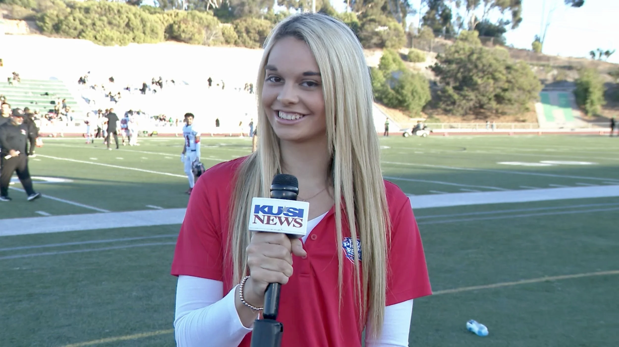 A young woman with long blonde hair, smiling and holding a microphone with a KUSI News logo, standing on a football field with players and spectators in the background.