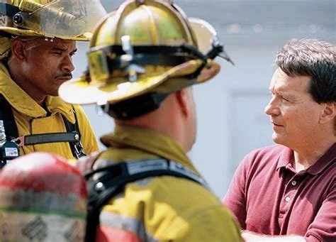A man in a maroon polo shirt talking to two firefighters in gear and helmets.