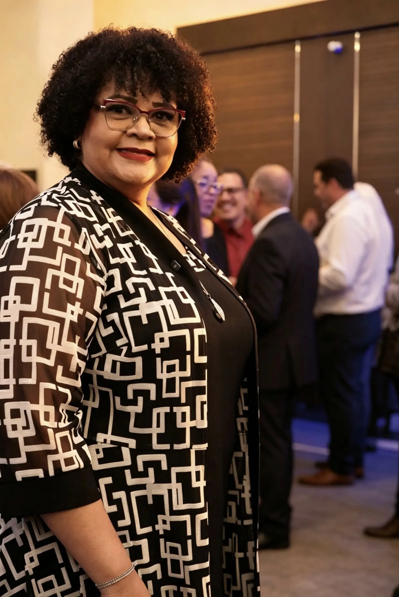 Una mujer con gafas y cabello rizado en un evento social, vestida con un abrigo de patrón geométrico en blanco y negro, en un ambiente con varias personas conversando al fondo.