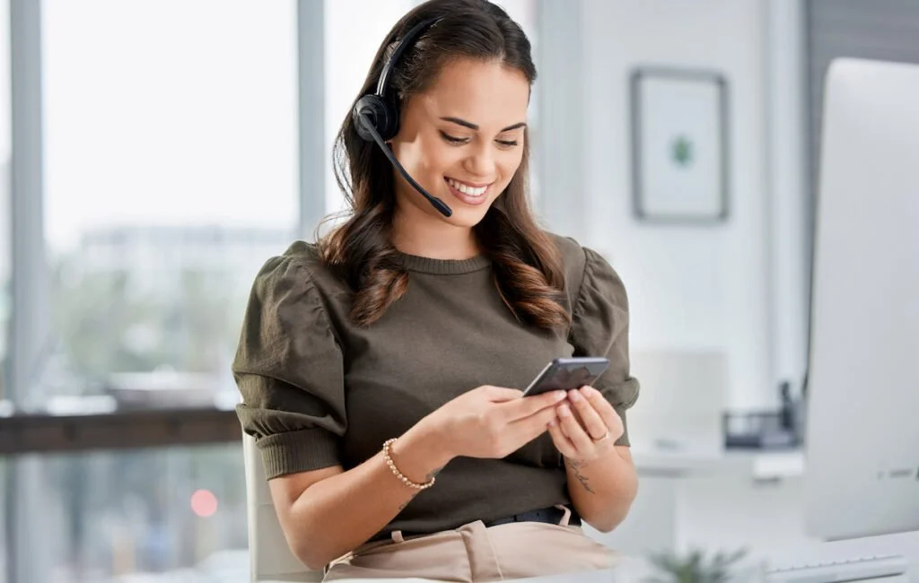 Mujer sonriente con auriculares de diadema y blusa gris, usando un teléfono móvil en una oficina moderna.
