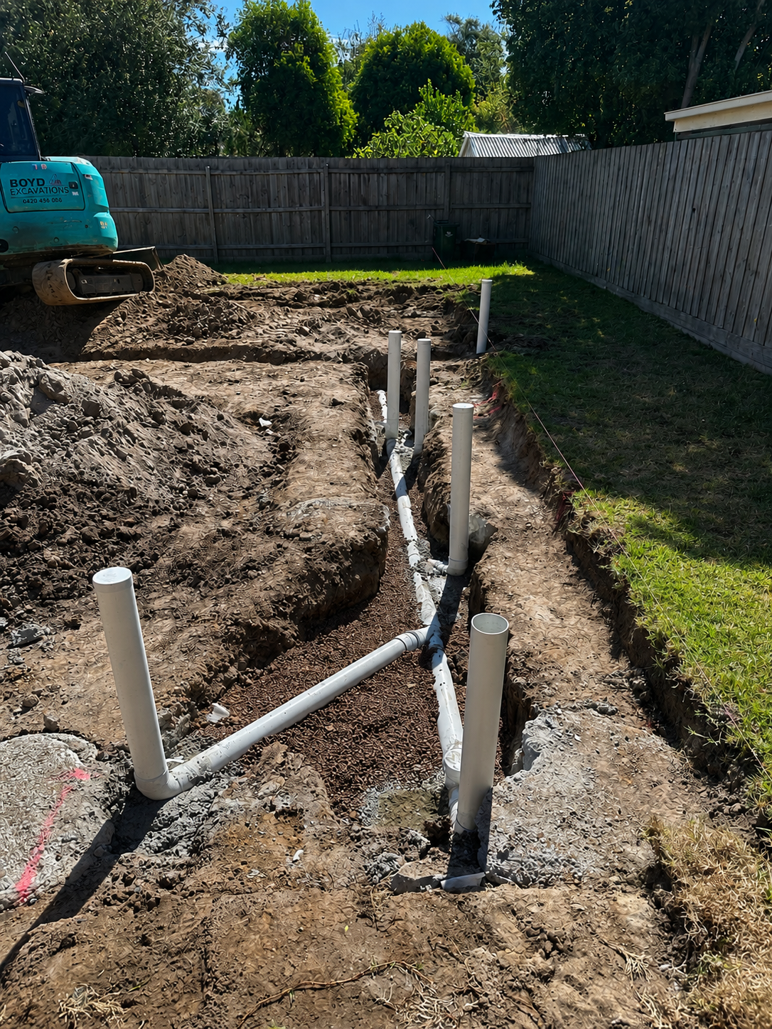 Underground plumbing installation with PVC pipes in a backyard dugout trench, surrounded by dirt and grass, with a wooden fence and trees in the background.