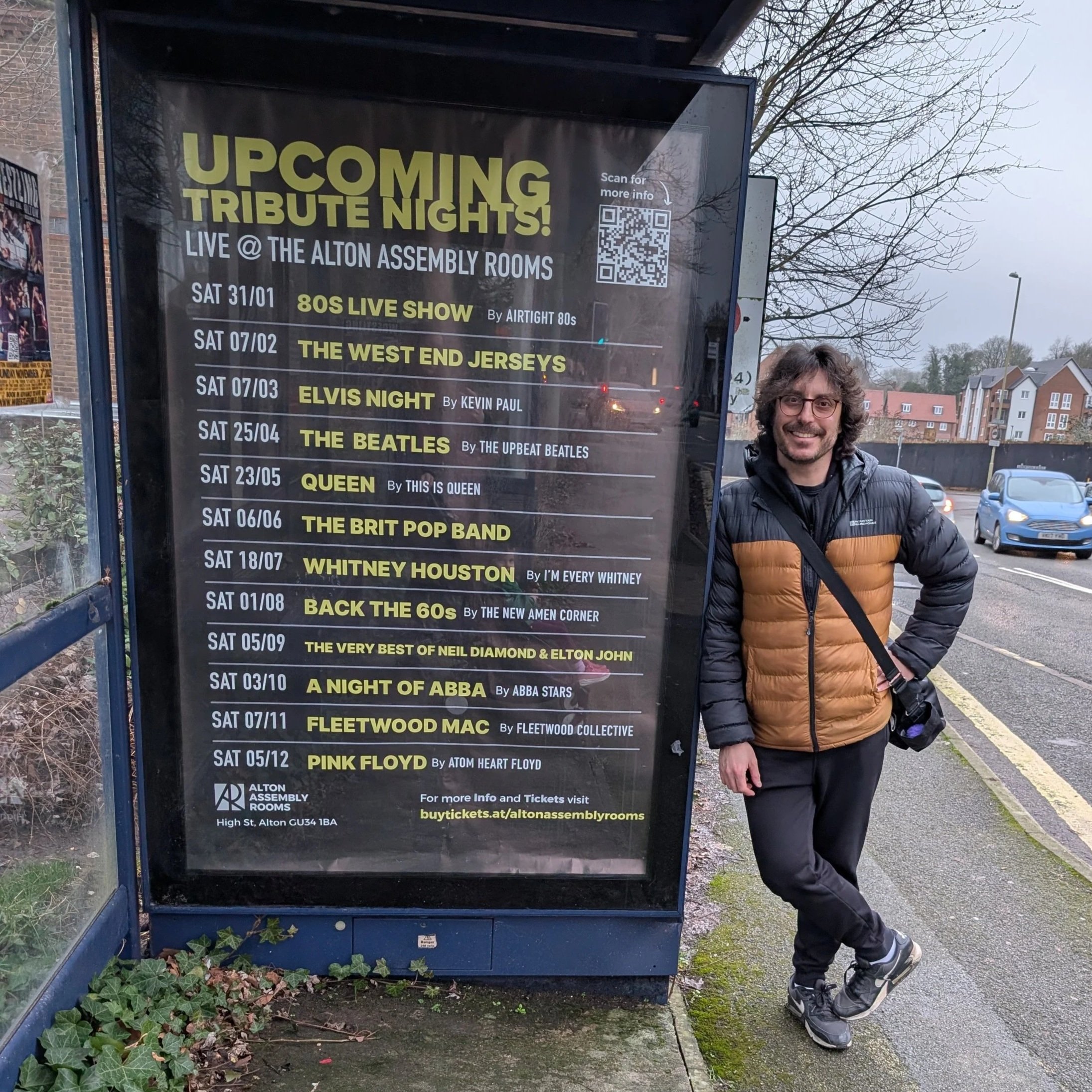A man with glasses and curly hair standing next to a bus stop display showing upcoming tribute nights at the Alton Assembly Rooms, outdoors with residential buildings and leafless trees in the background.