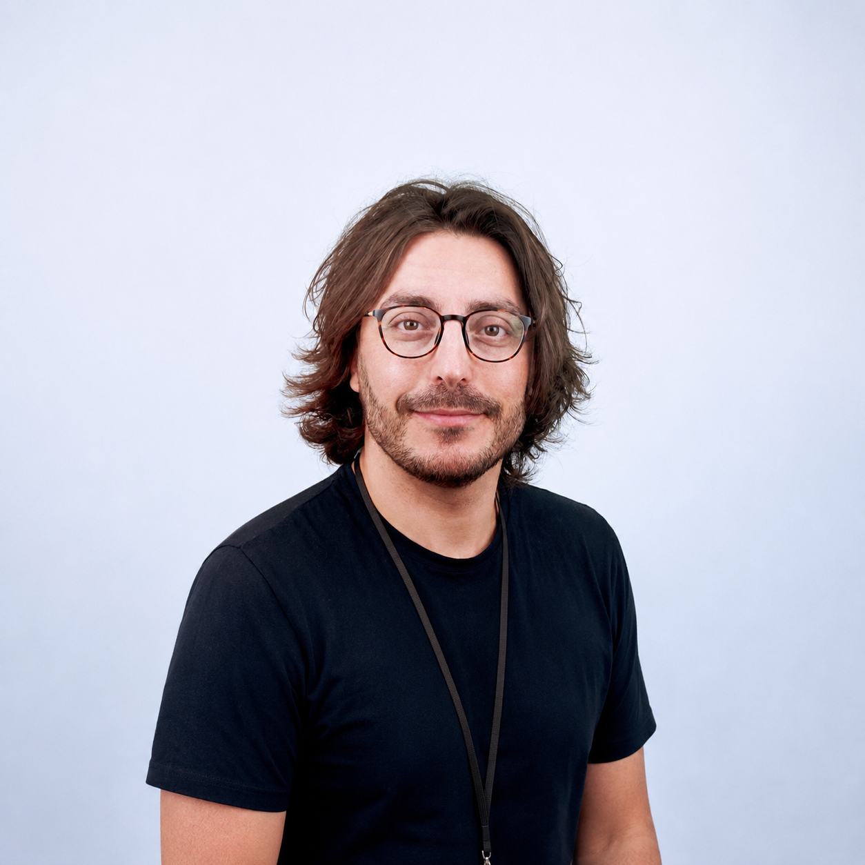 Portrait of a young man with shoulder-length brown hair, glasses, and a beard, wearing a black t-shirt and a lanyard against a plain light gray background.